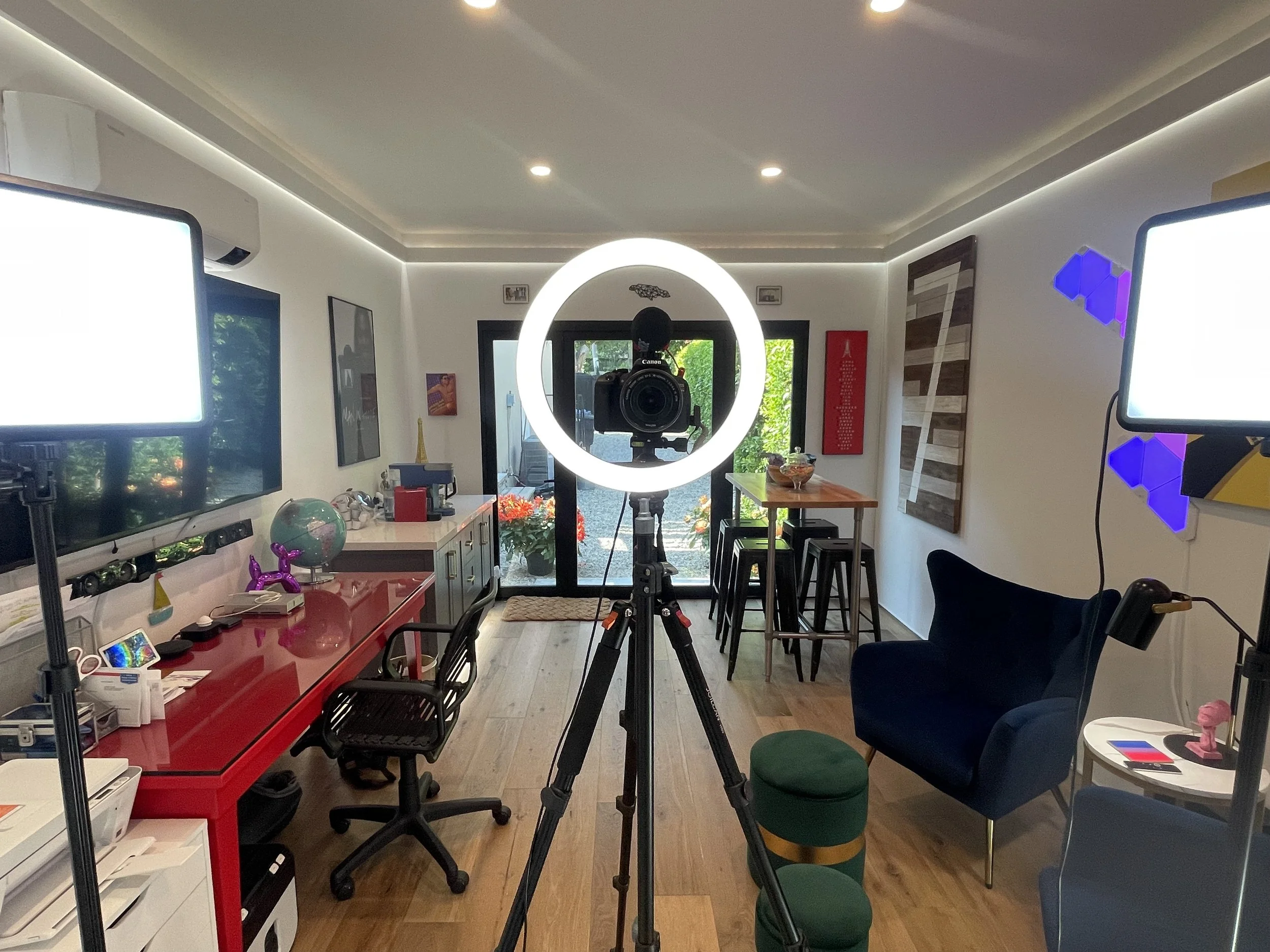 Home studio setup with camera on tripod centered, two computer monitors on left and right, a red desk, a black chair, a navy armchair, and a small round side table, with decorations and plants near a glass door.