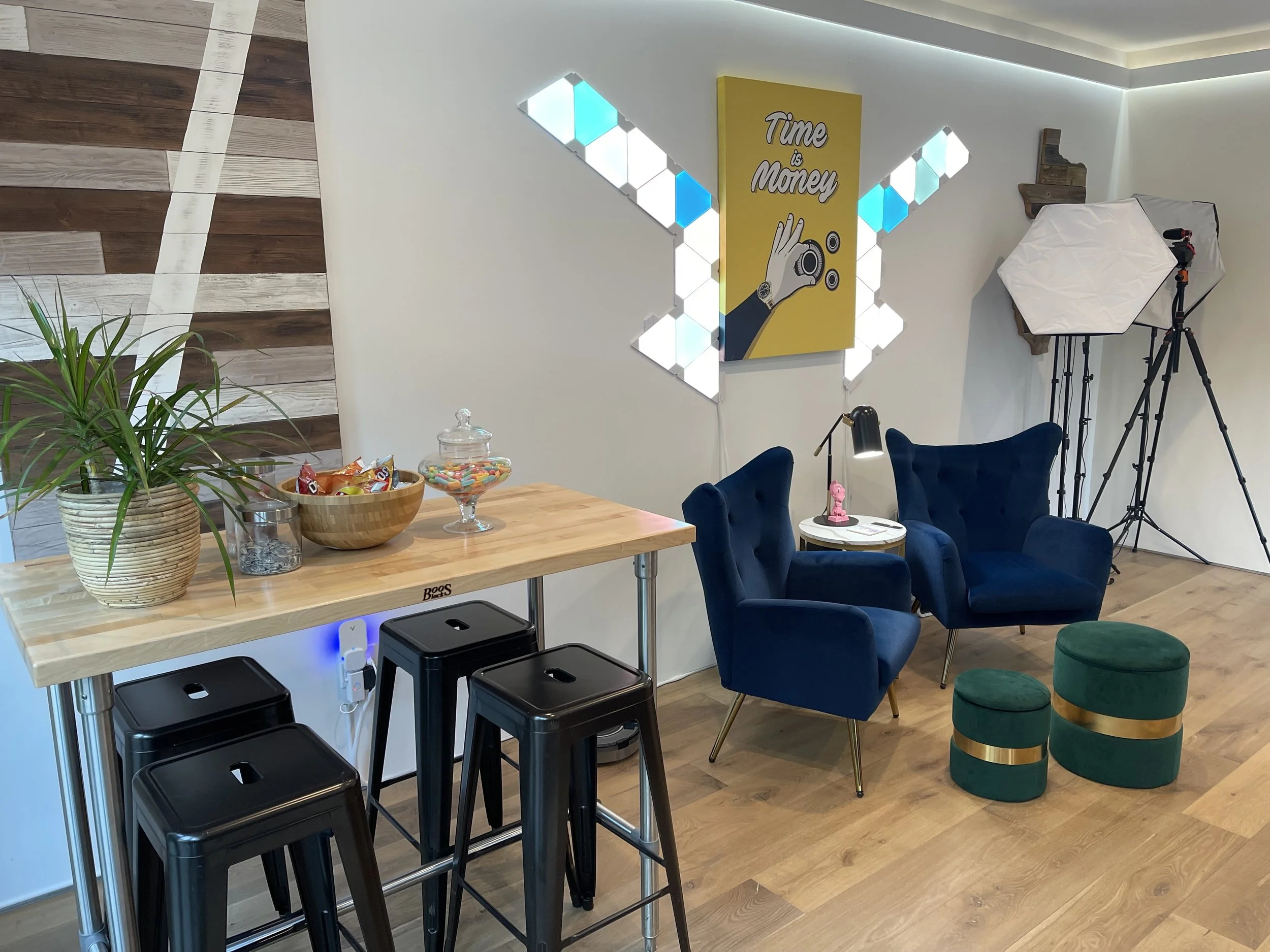 A modern studio space with a wooden counter, black bar stools, potted plant, bowl of candy, and glass jar on top. There are two blue velvet armchairs, a small round white table, and two green ottomans with gold bands. Photography equipment including a tripod and a softbox light are set up in the corner. A yellow wall art piece reads 'Time is Money' with a hand holding a camera.