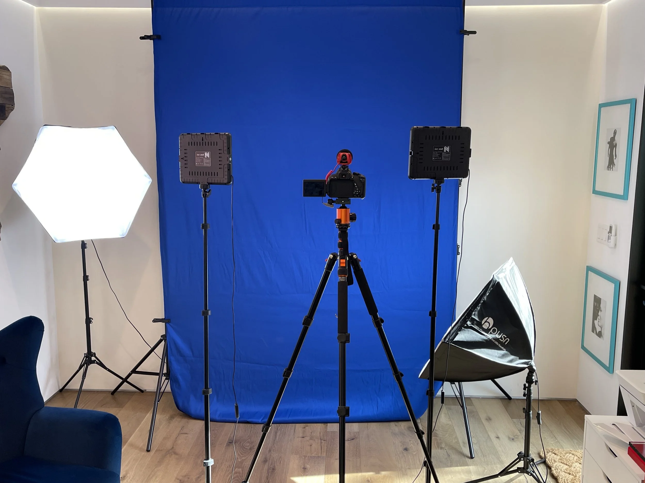 Home photography studio setup with a blue backdrop, three lighting panels, a camera on a tripod, and photography equipment in a living room.