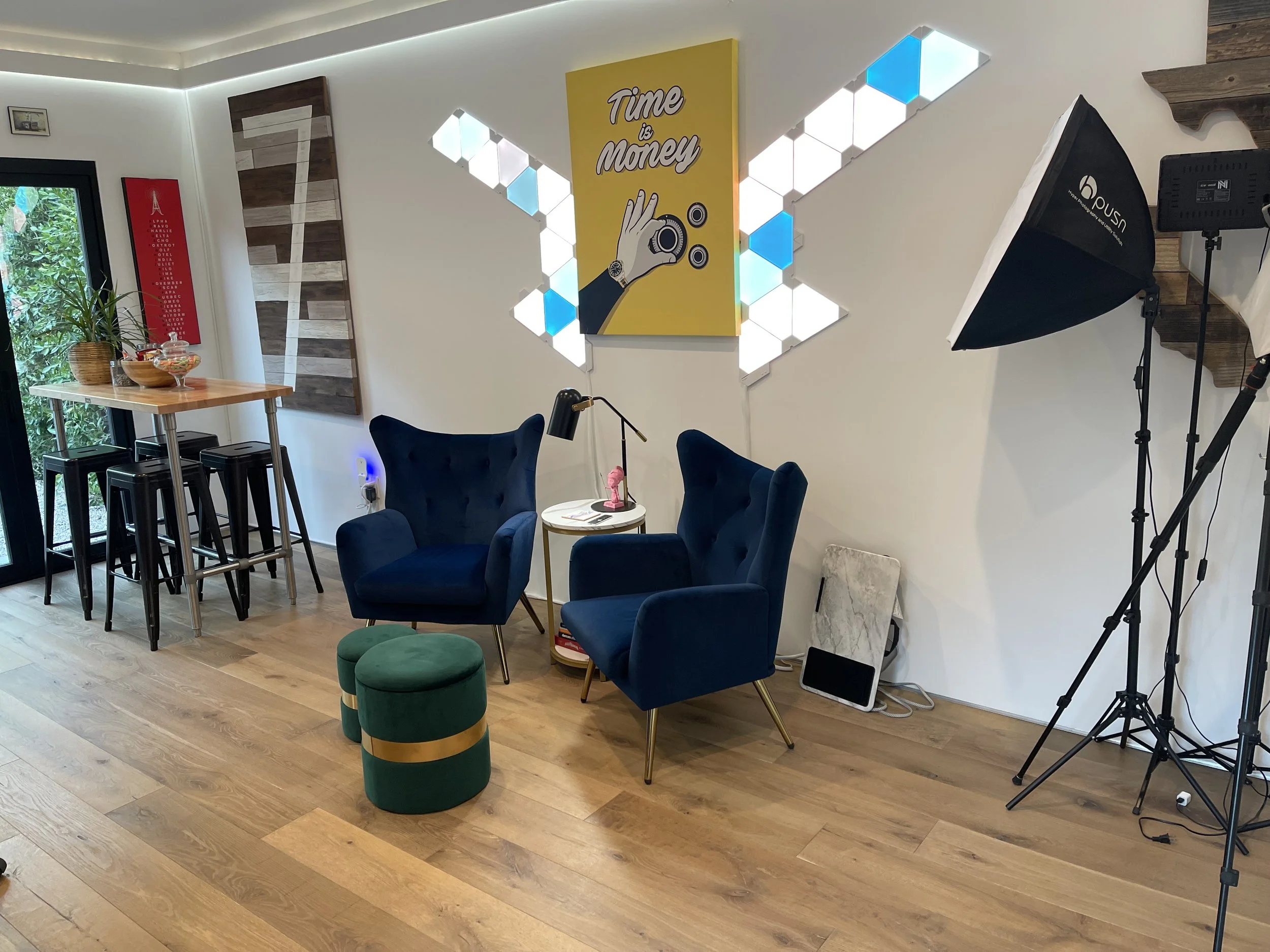 A cozy room set up for a photo or video shoot with two blue armchairs, a small side table with a pink decorative item and a black desk lamp, a green and gold round ottoman, photography lighting equipment, and decorative wall art including a yellow po