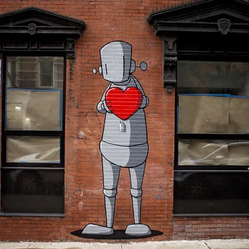 Street art mural of a robot holding a red heart on a brick wall between two windows.