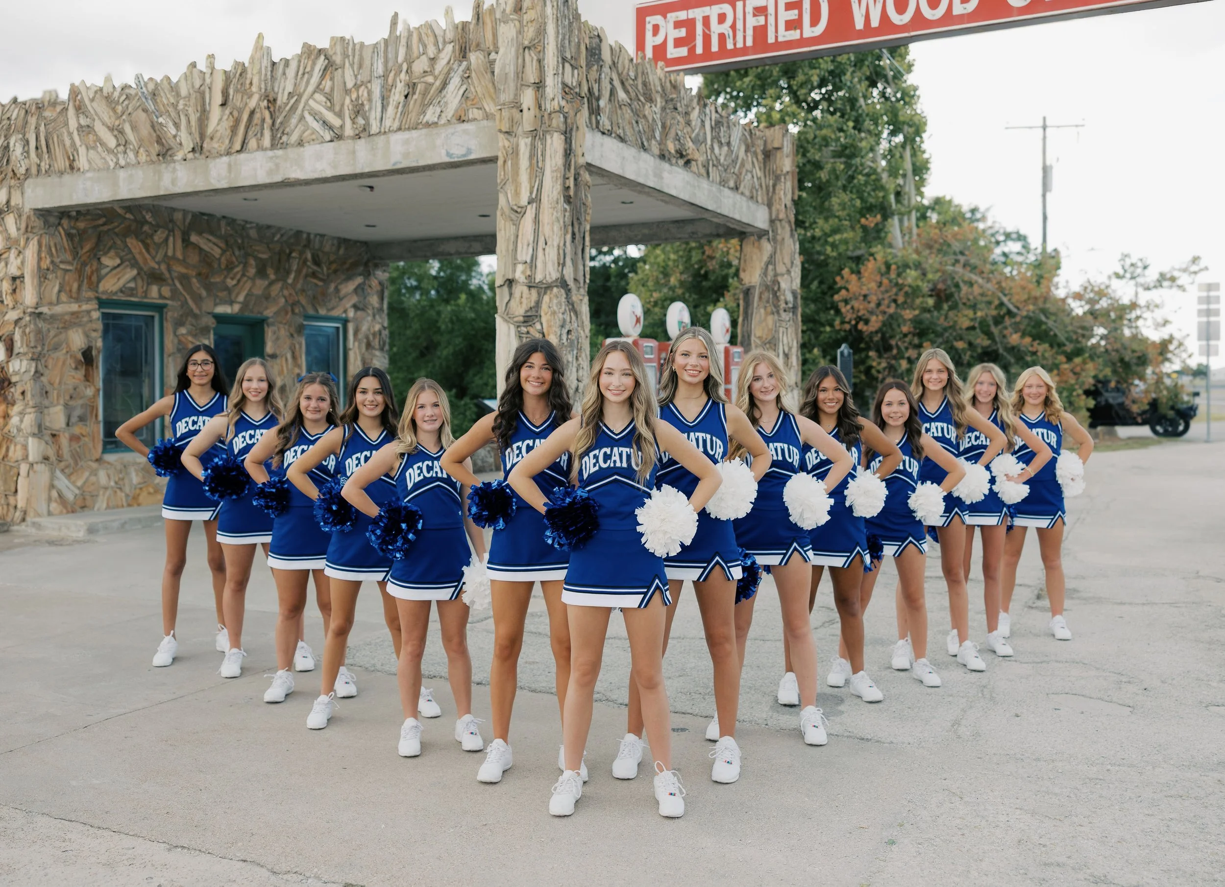 Group of teenage cheerleaders in blue uniforms standing on a football field, holding hands in a line, with a scoreboard and school buildings in the background.