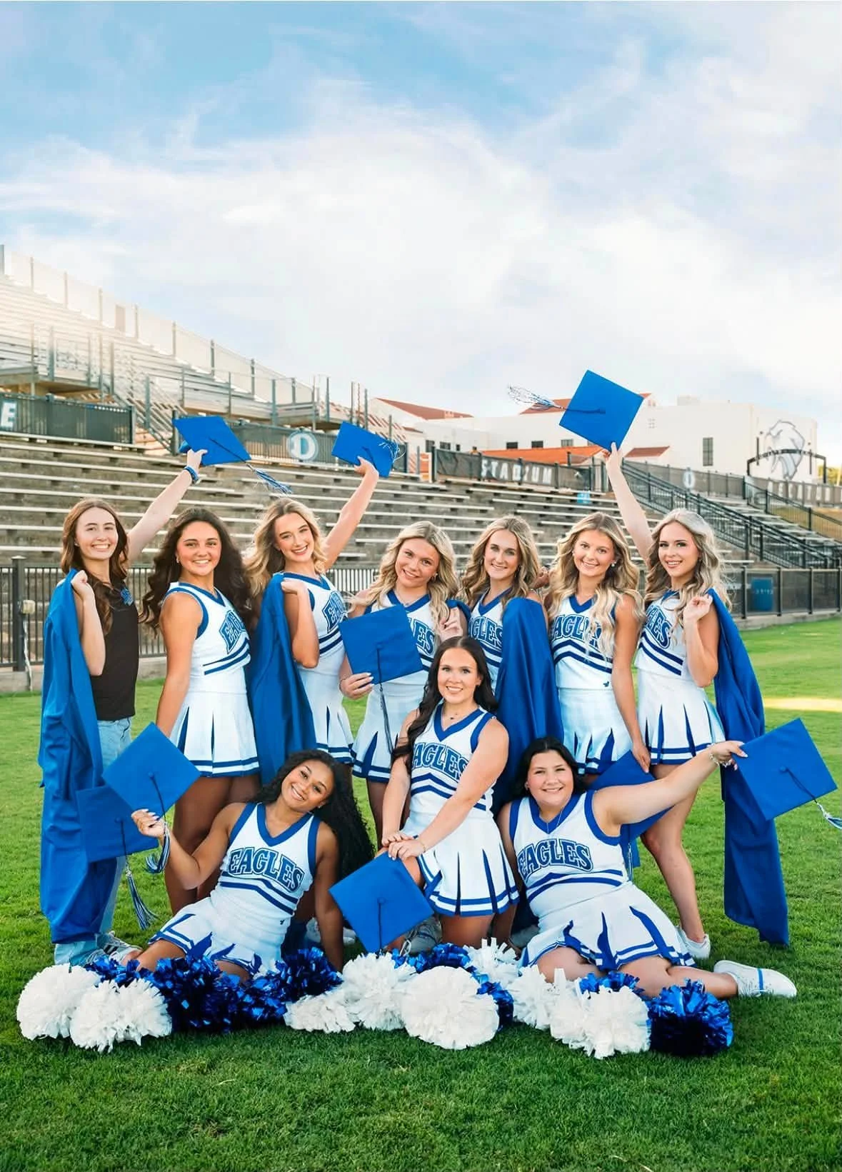 Group of cheerleaders in blue and white uniforms standing on a football field with a stadium and bleachers in the background, along with a person dressed as an eagle mascot.