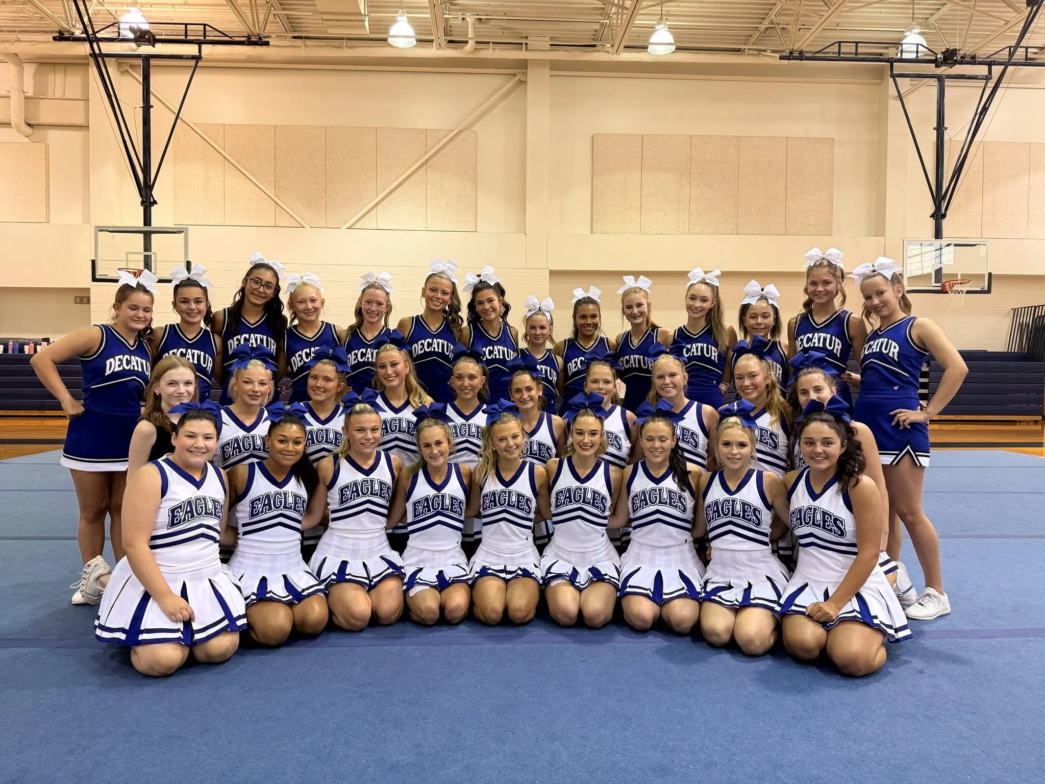 A group of 14 cheerleaders wearing blue uniforms with 'Decatur' written on the front, standing outdoors on a street, posing with their hands on their hips and holding pom-poms, smiling at the camera.