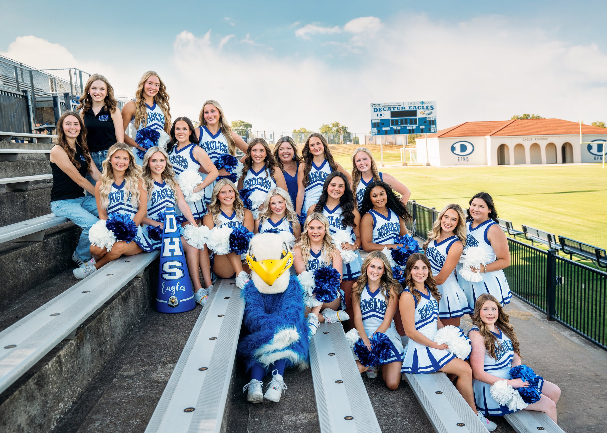 A cheerleading team in blue and white uniforms posing on a football field with a mascot and cheerleaders in front of empty bleachers at a stadium during the daytime.