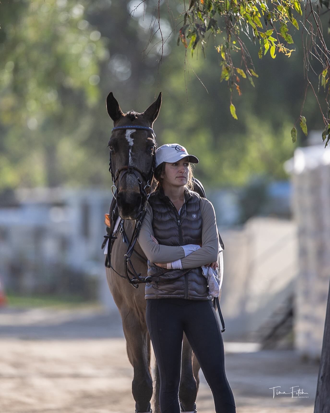 A woman stands with her arms crossed next to a brown horse on a trail in a wooded area. The woman is wearing a white cap, a gray vest, and black athletic pants, while the horse is fitted with riding gear. The background is blurred with green and gray