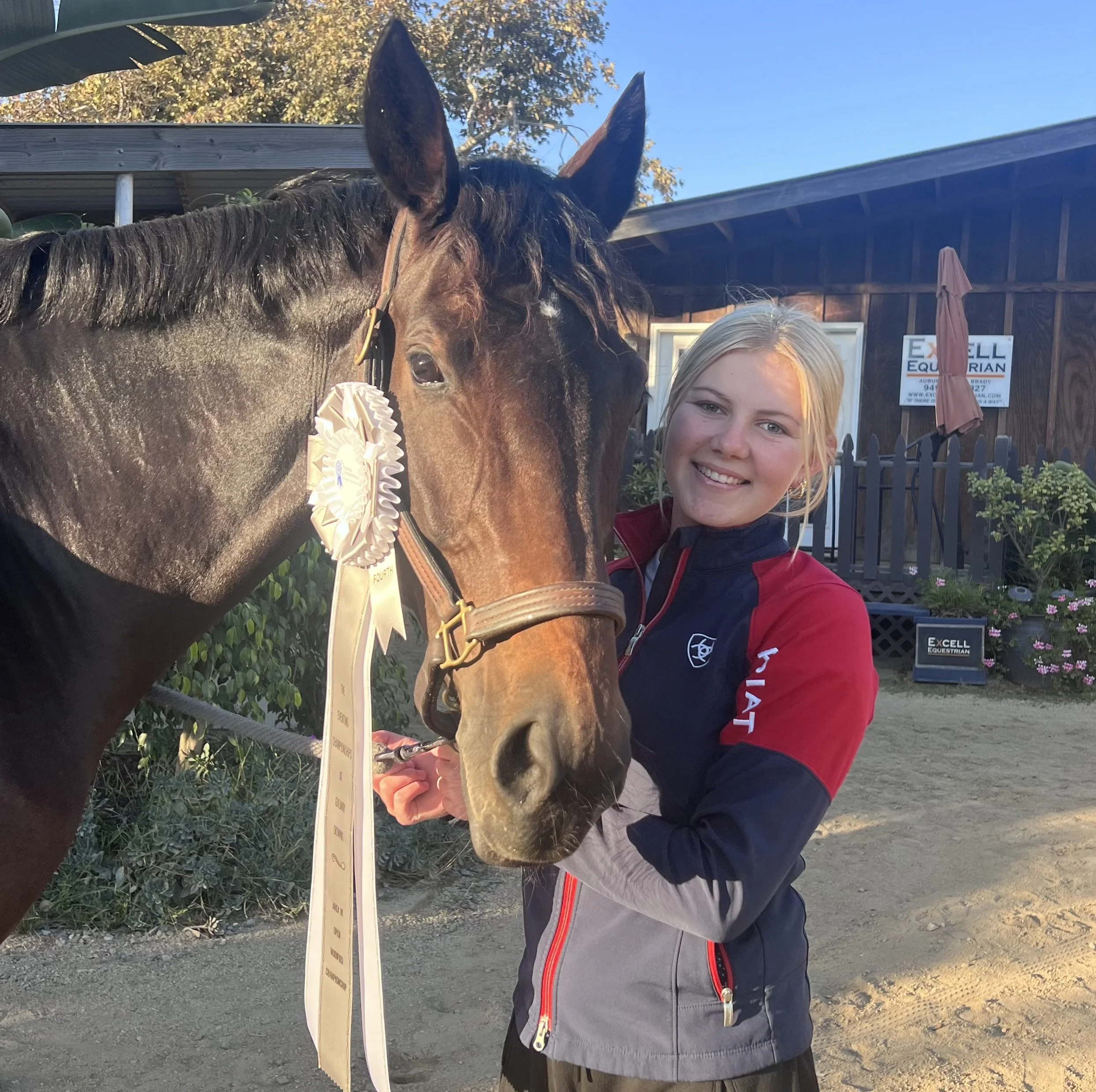 A young girl with blonde hair smiling at the camera, standing outdoors next to a brown horse with a white stripe on its face. The horse is adorned with a ribbon and a rosette, indicating it has won an award. Behind them, there is a wooden fence, a si