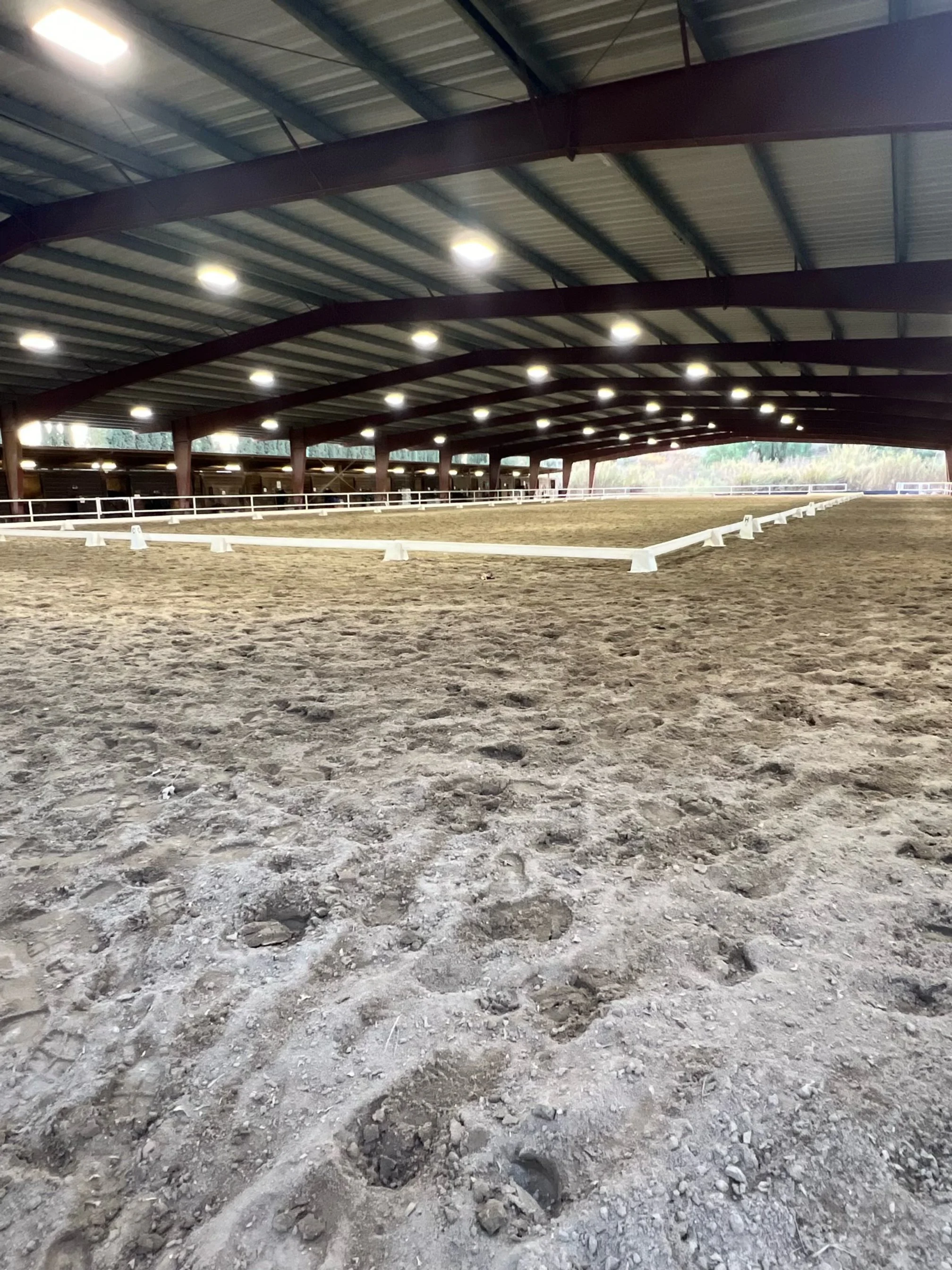 Indoor horse riding arena with a dirt floor, white fencing, and a metal roof supported by red beams, illuminated by overhead lights.
