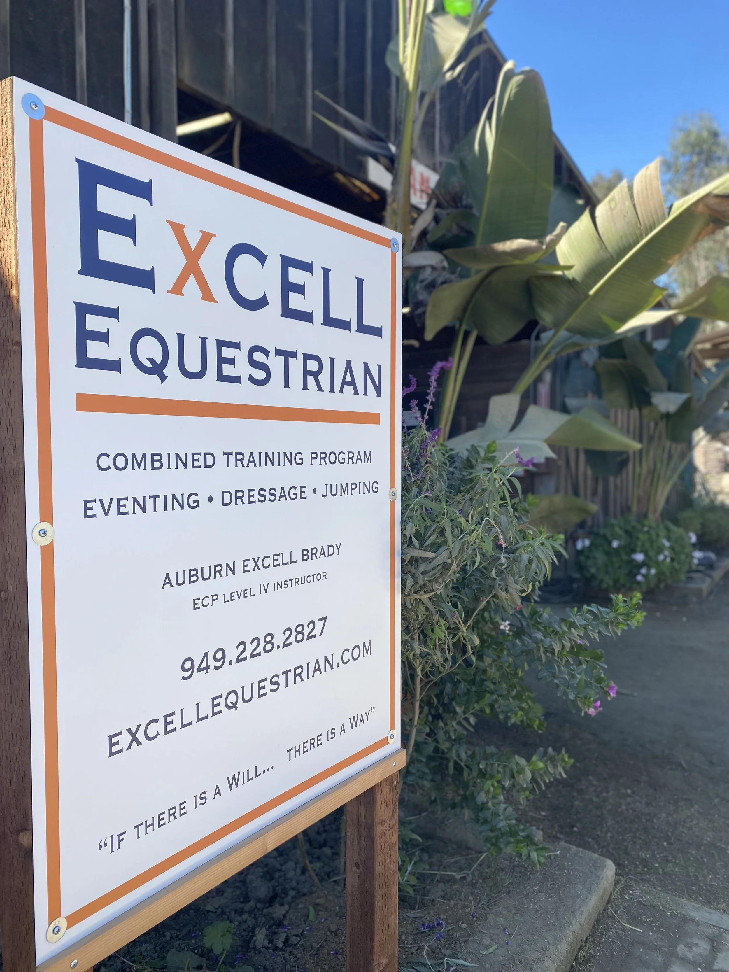 A sign for a combined training program in equestrian activities, including eventing, dressage, jumping, and more, displayed outdoors next to lush green plants under a clear blue sky.