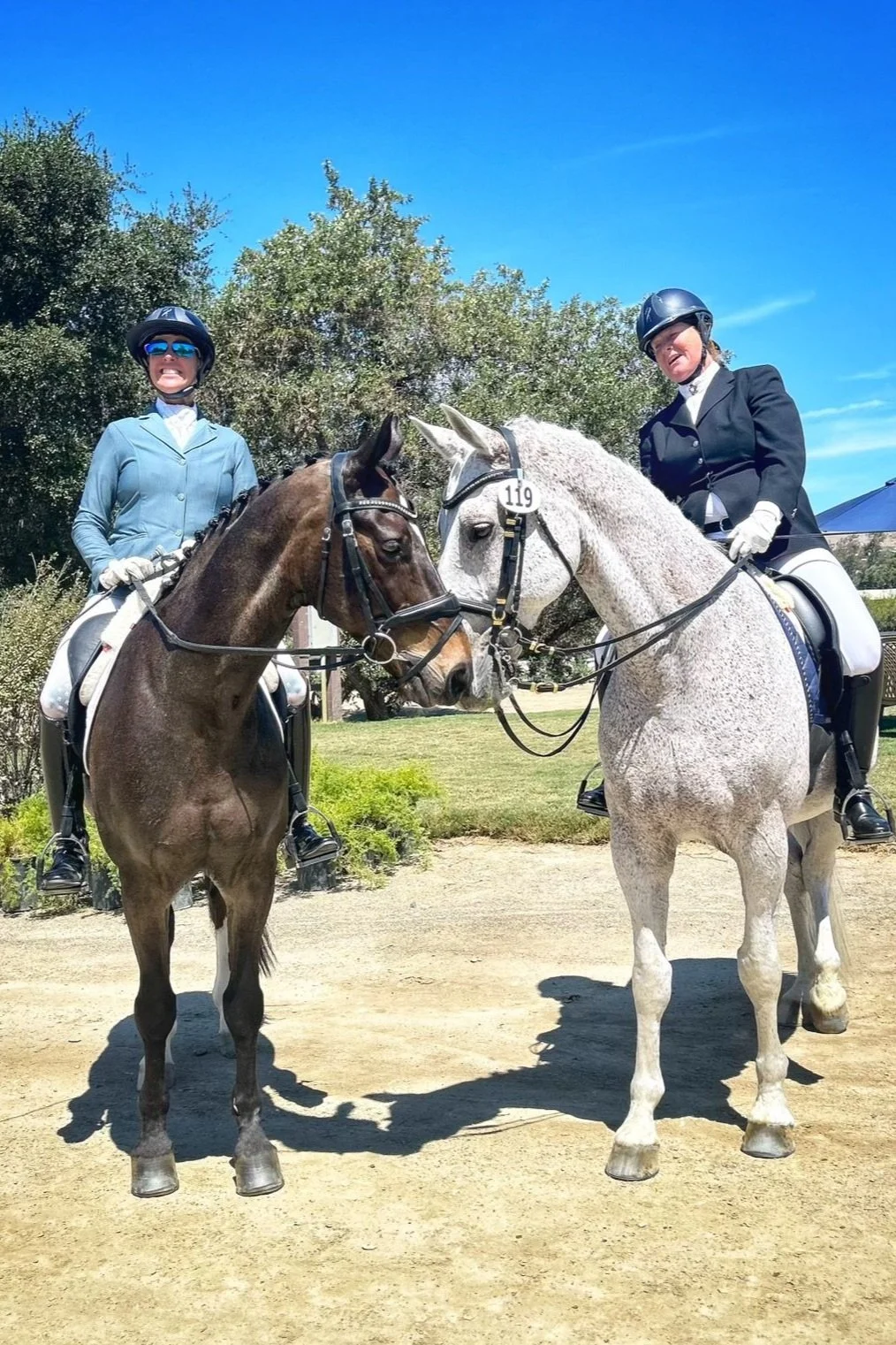 Two women in equestrian attire sitting on horses, touching foreheads, outdoors on a sunny day with trees and blue sky.