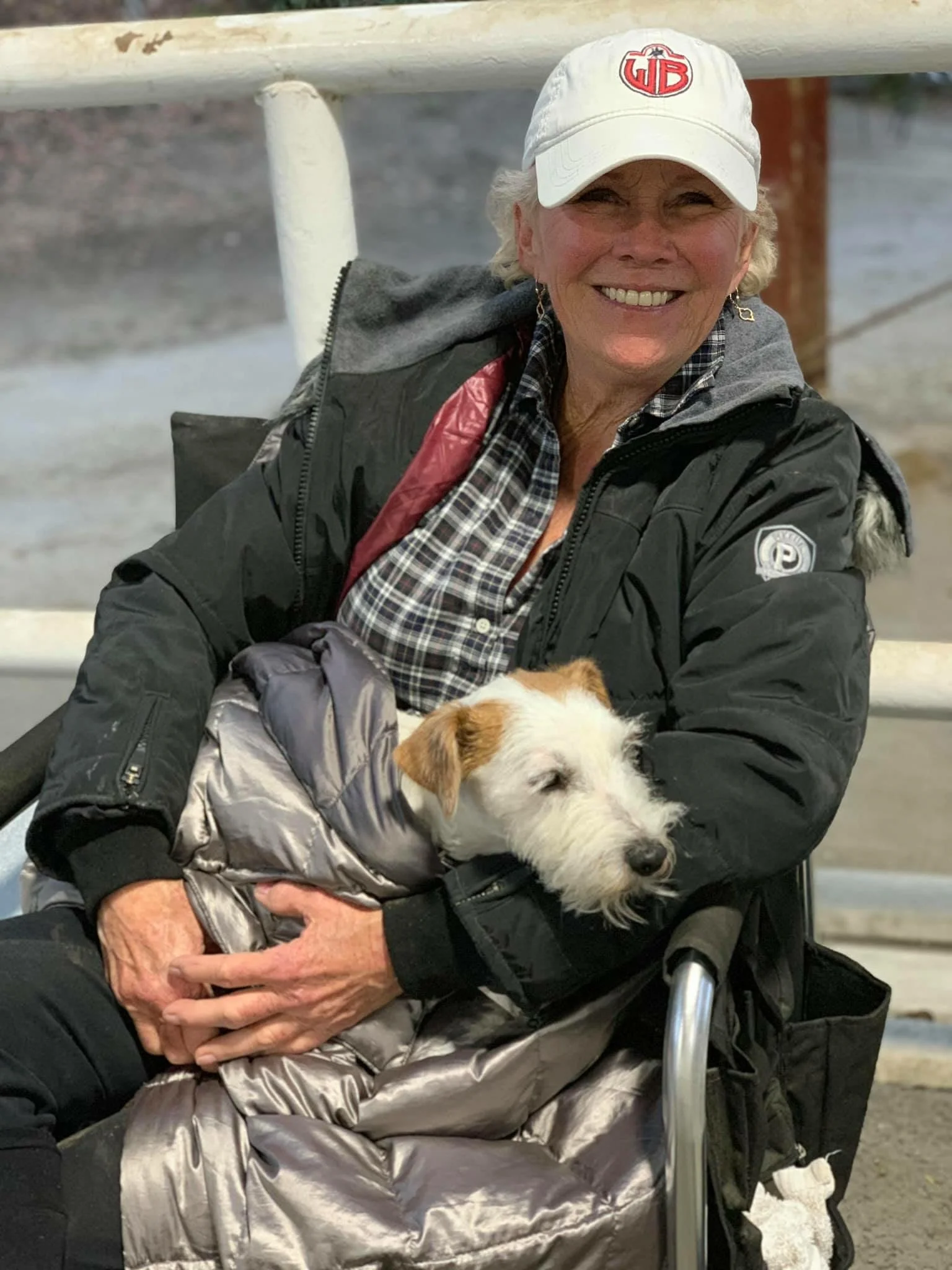 A smiling elderly woman with white hair wearing a white cap, plaid shirt, and black jacket, sitting in a wheelchair, holding a small white and brown dog.