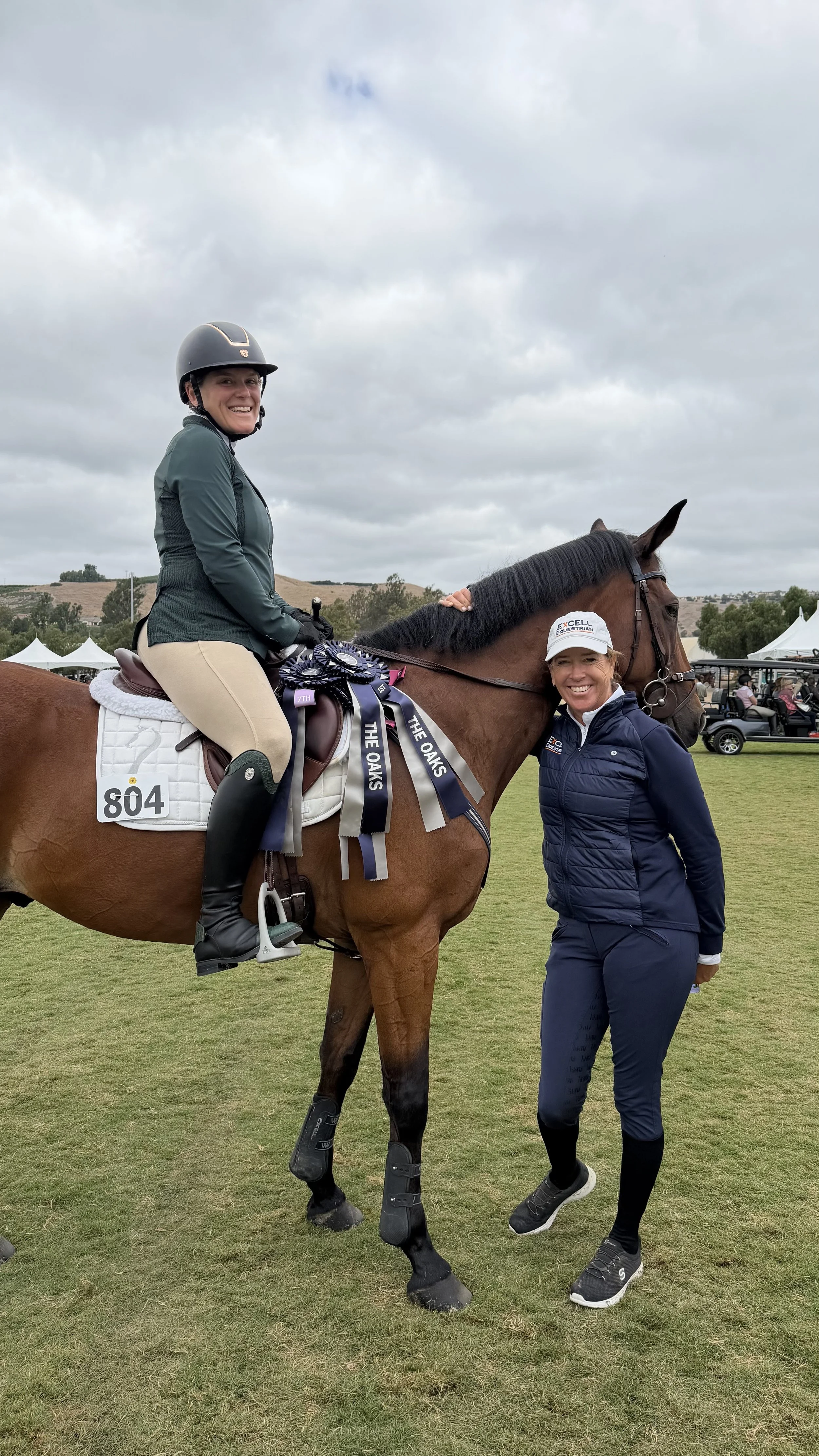 A woman riding a horse with ribbons on its saddle and a woman standing next to the horse smiling, in an outdoor equestrian event on a cloudy day.