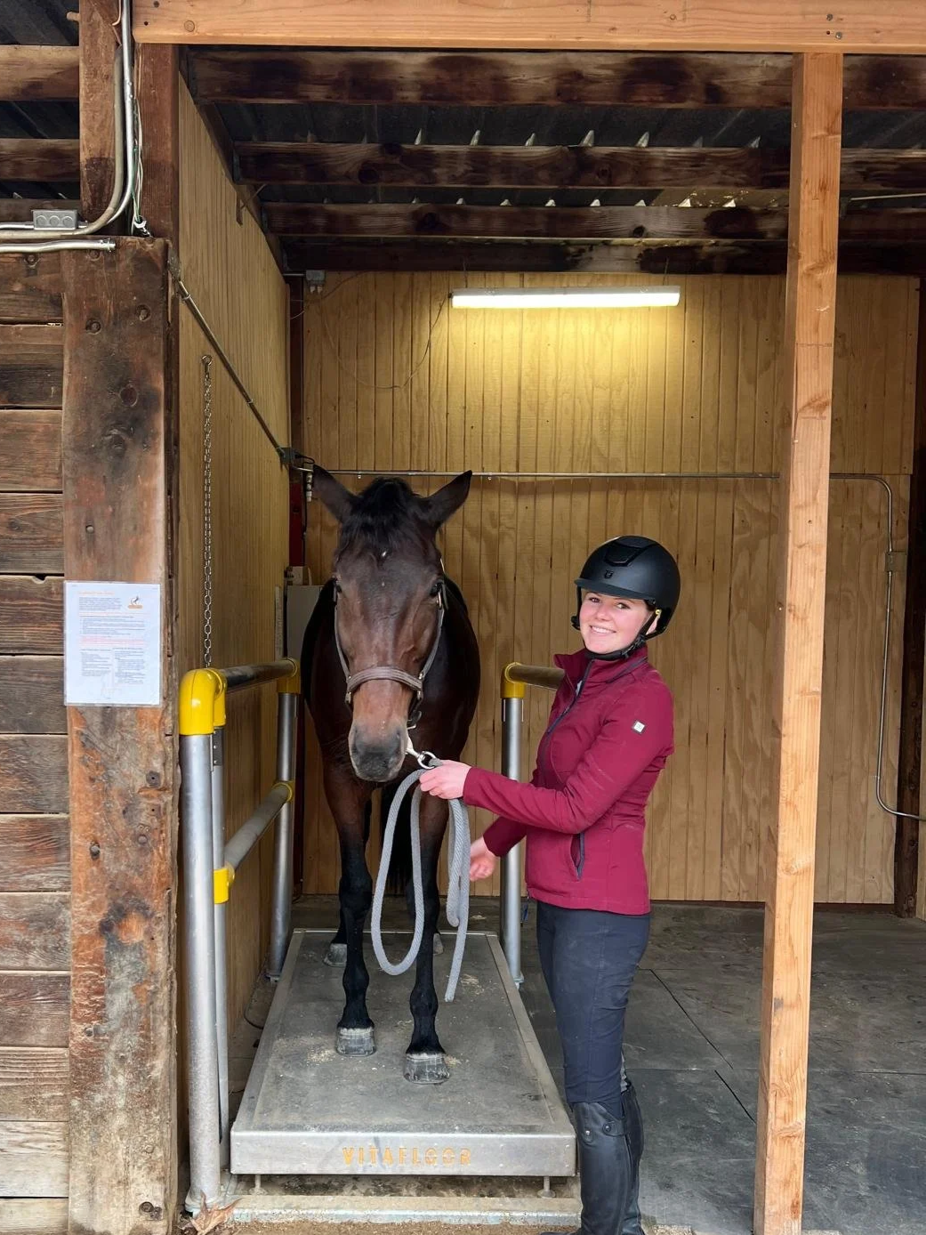 A woman in riding gear and a helmet standing beside a brown horse in a small wooden horse stall, holding the horse's lead rope and smiling.
