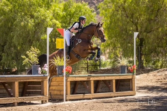Young female equestrian riding a brown horse jumping over an obstacle in a competition outdoors with trees and dirt ground.