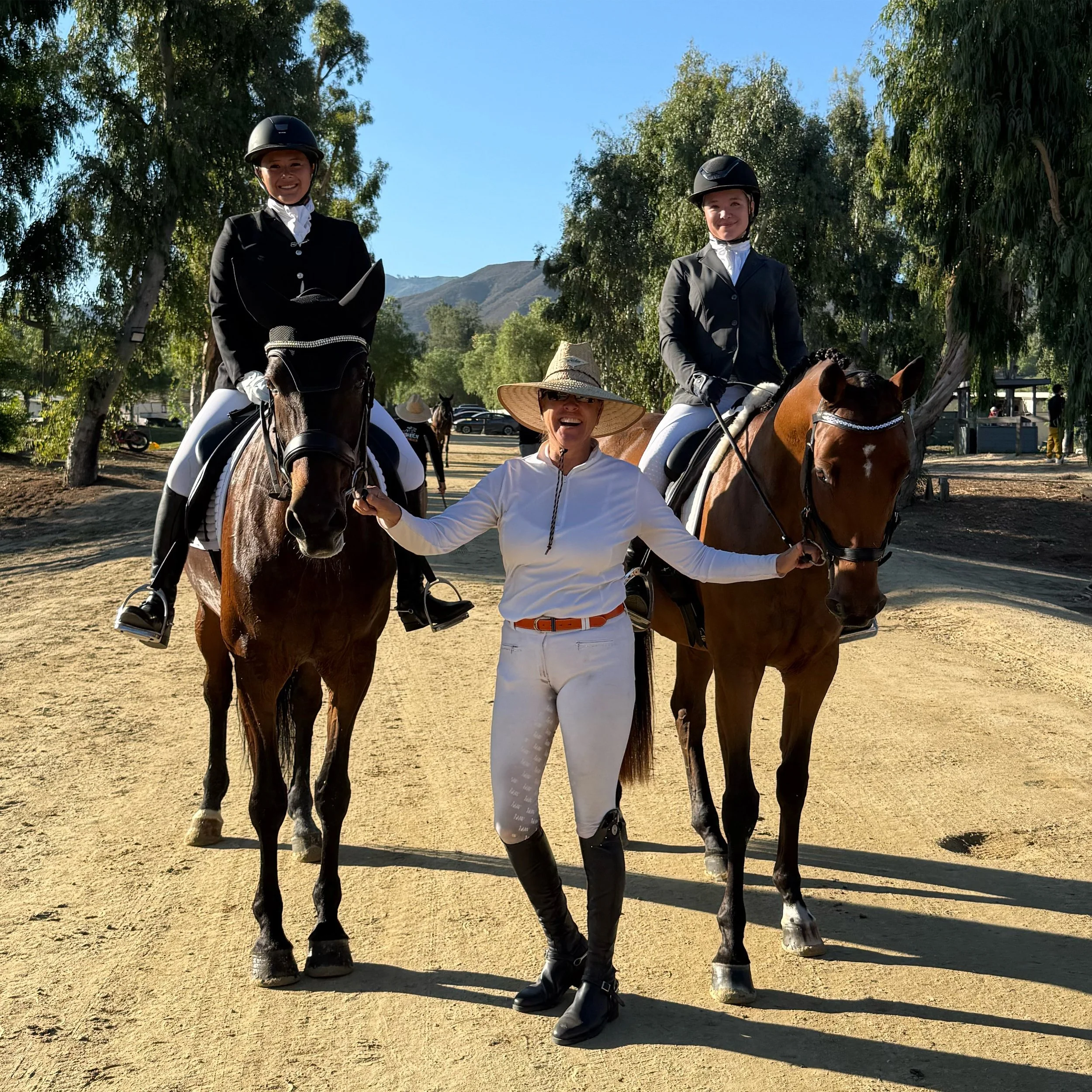 Three women riding horses outdoors near trees, with mountains and a clear blue sky in the background. One woman is holding a riding crop and smiling.