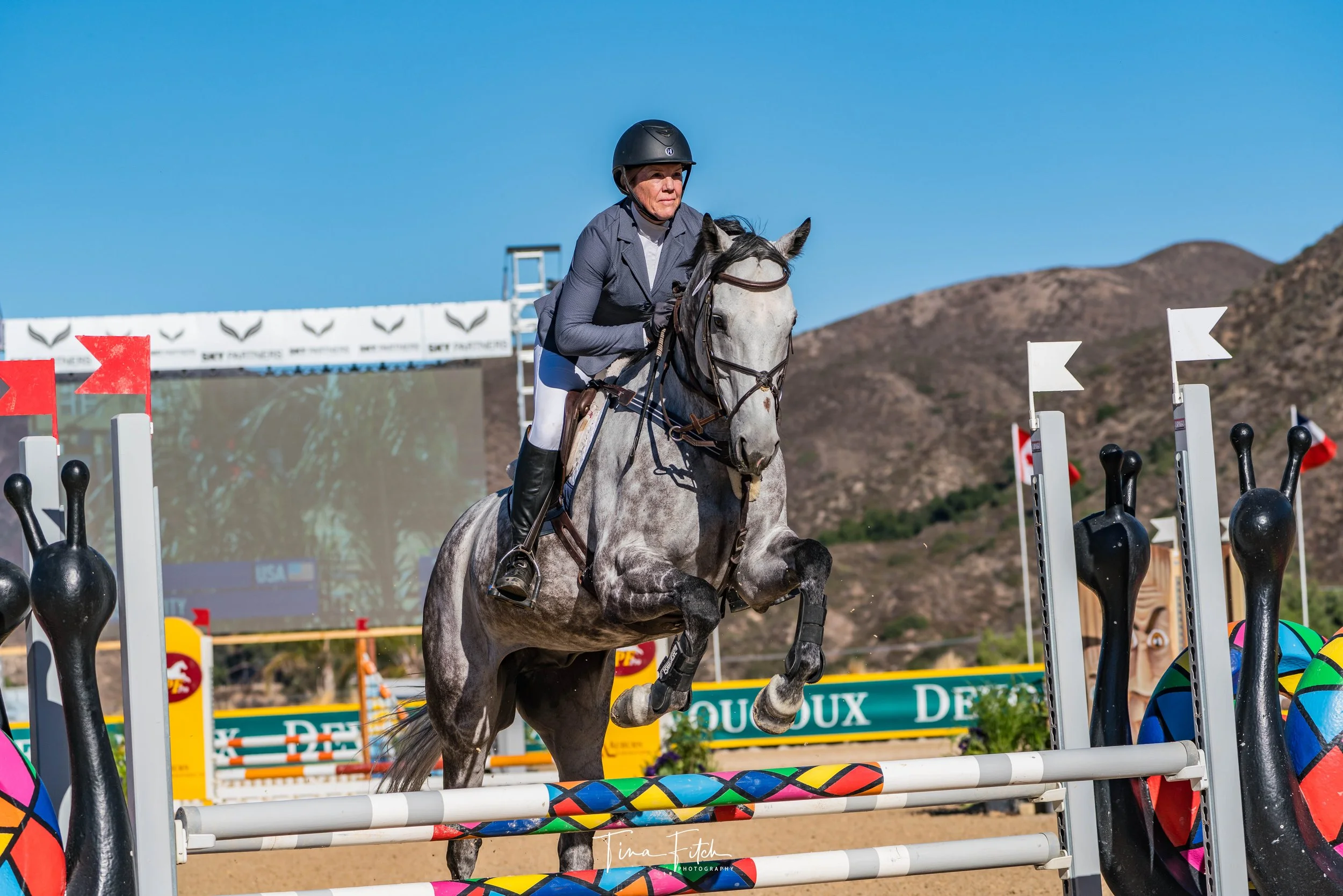 A person riding a gray horse over a show jumping obstacle on an outdoor equestrian course. The rider is wearing a helmet, a jacket, white riding pants, and boots. The background features a clear blue sky, hills, and promotional banners.
