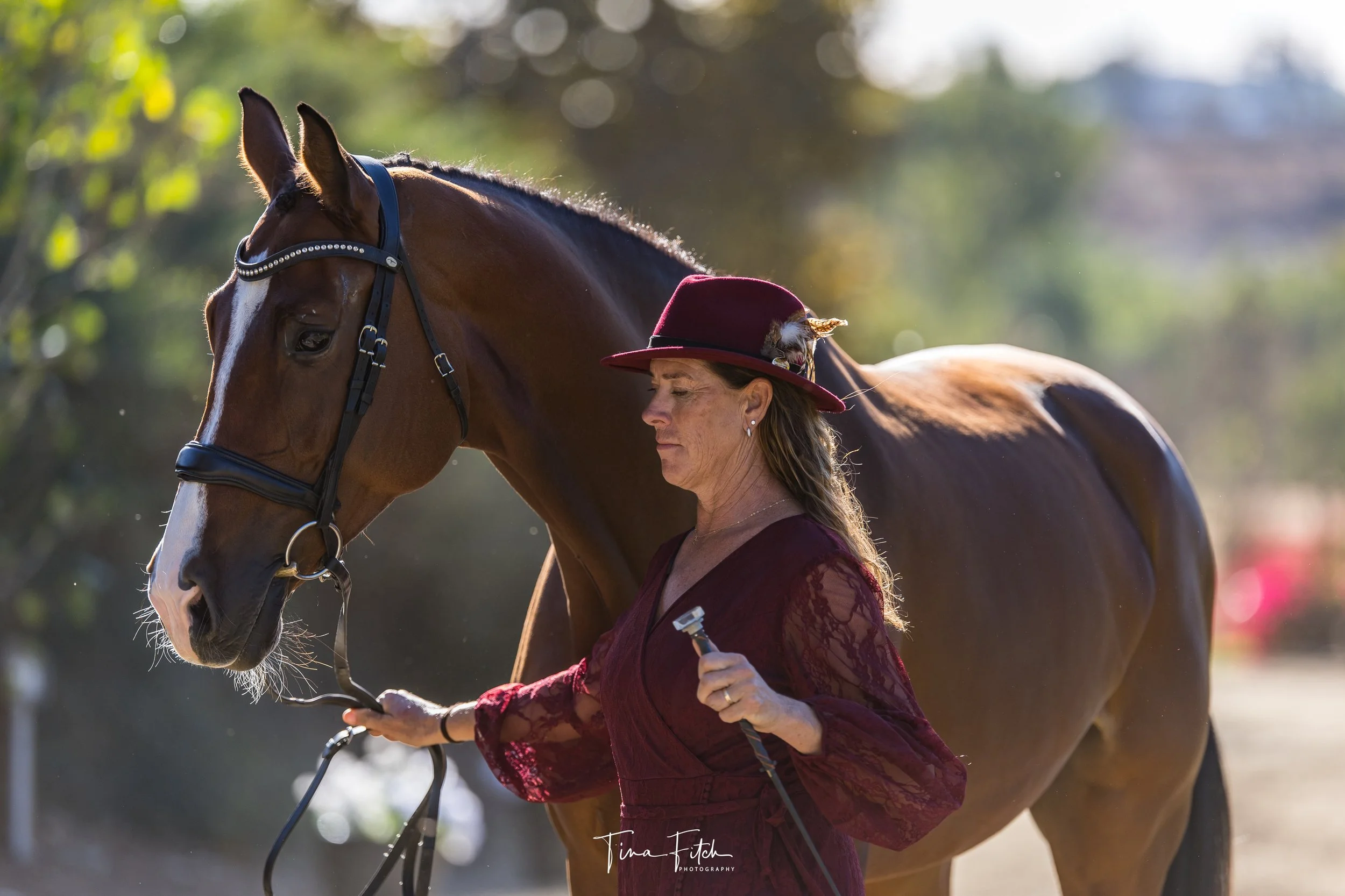A woman in a maroon dress and hat with a feather, standing beside a brown horse, gently holding the horse's bridle as the horse looks down. The background is blurred with trees and sunlight.