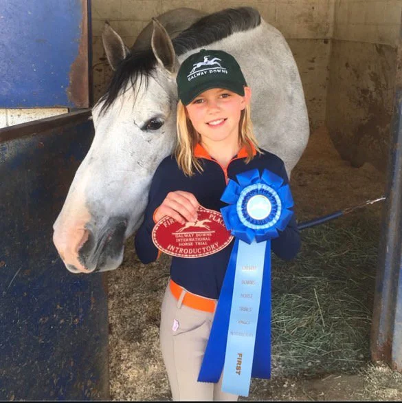 A young girl with a big smile, wearing a black cap and a dark blue jacket, stands inside a stable holding a red award ribbon and a blue first-place rosette, with a gray and white horse beside her.