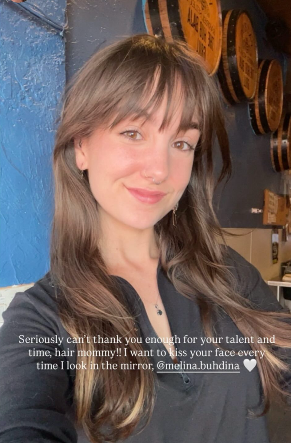 A young woman with long brown hair, smiling, wearing a nose ring, earrings, a black top, and a necklace. She is in a room with blue-painted walls and wooden barrels in the background.
