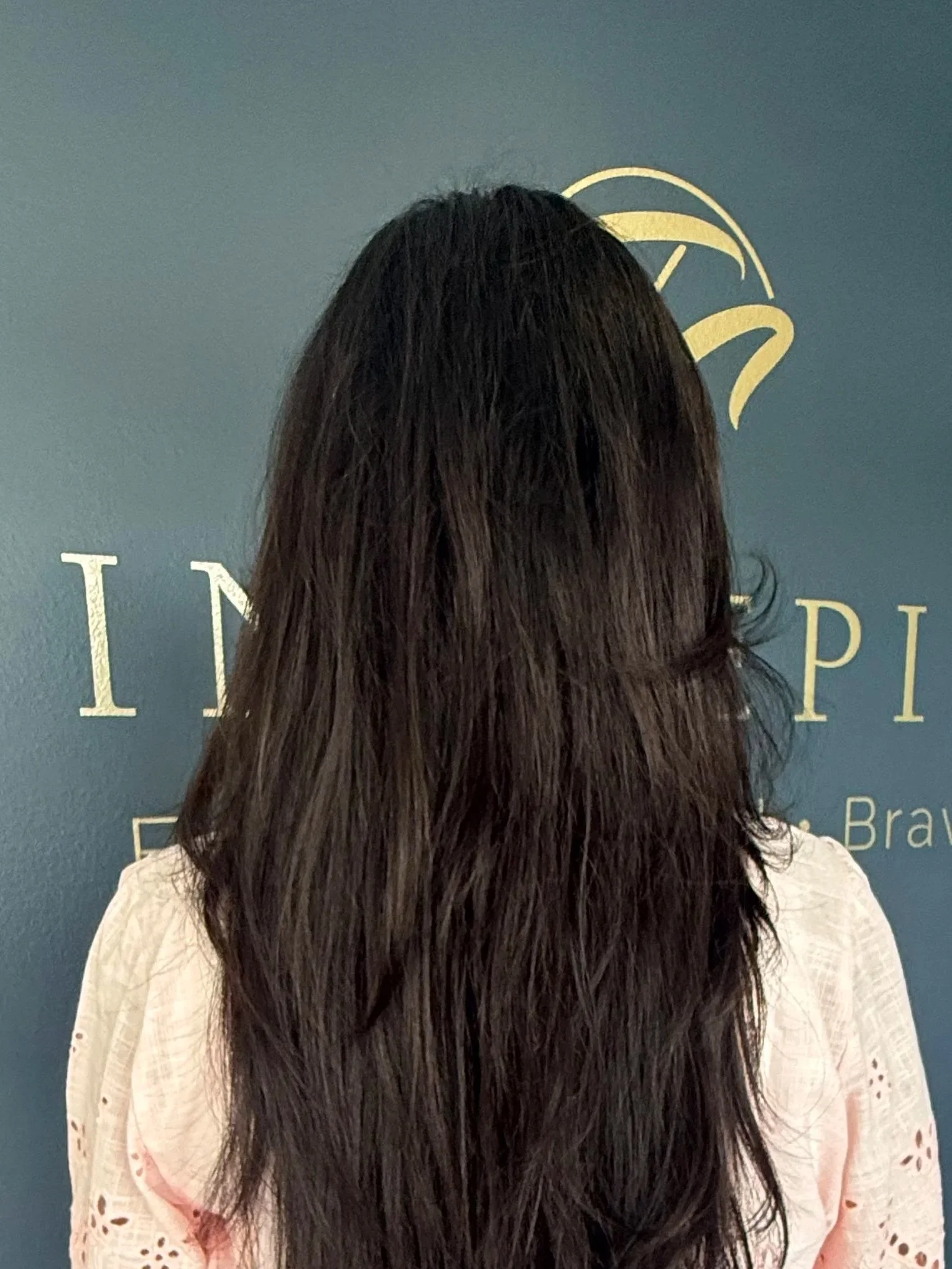 A woman with long dark hair standing in front of a blue wall with gold-colored lettering and logo, wearing a white lace-patterned shirt.