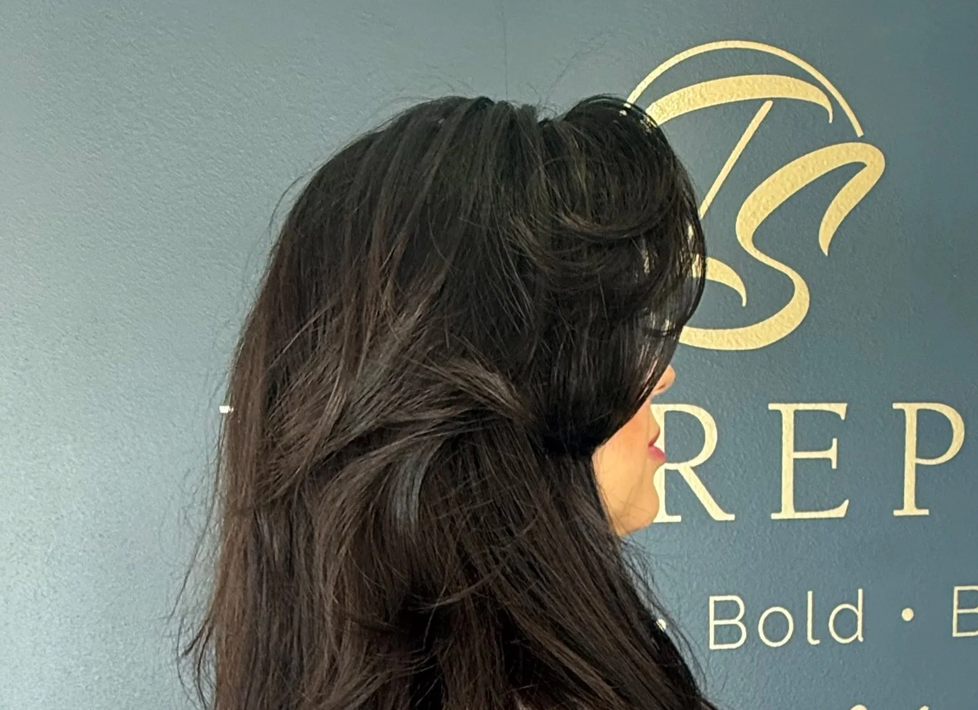 A woman with dark brown, wavy hair looking to the right, standing in front of a blue wall with a gold logo and text.