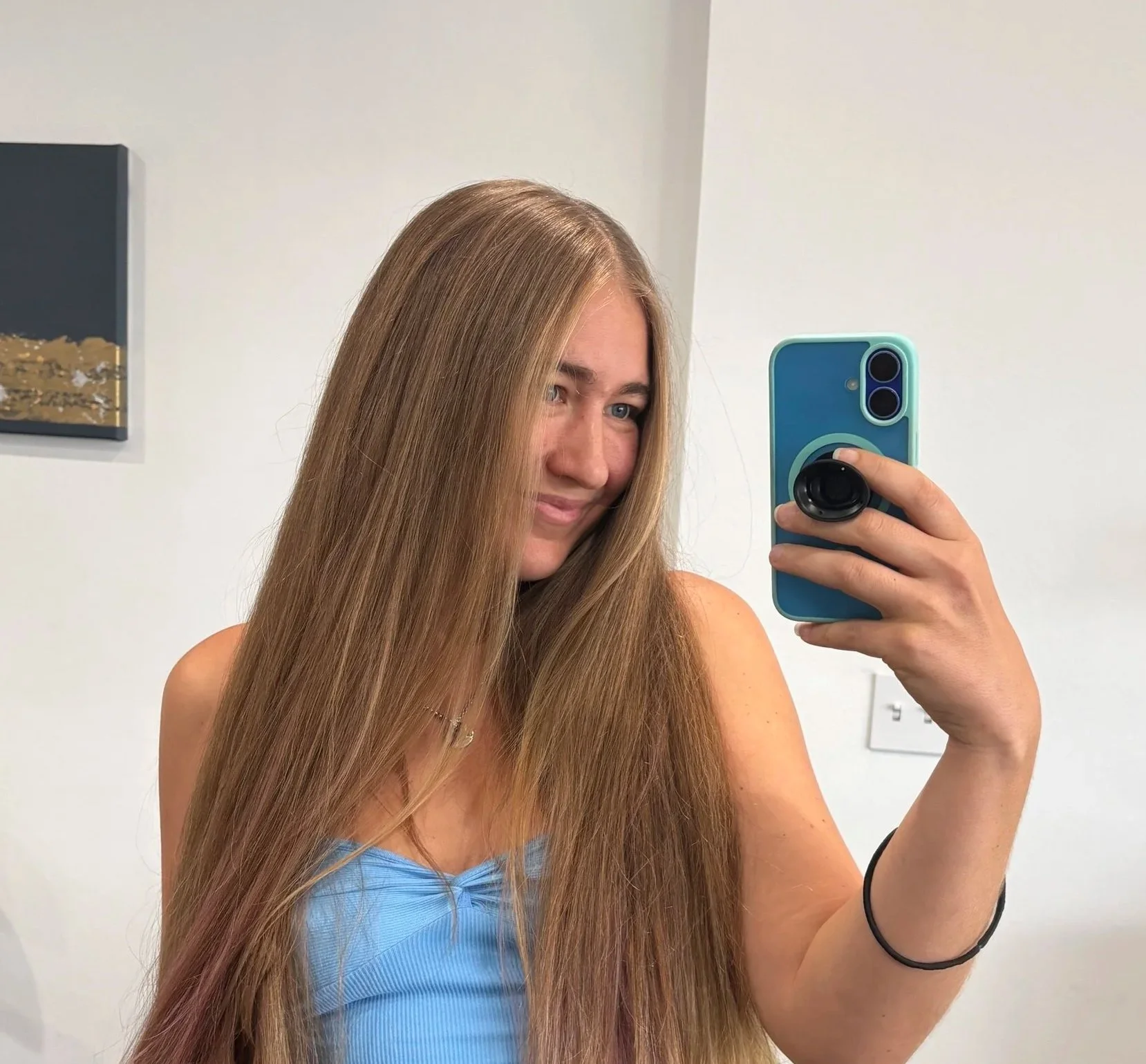 A woman with long, straight brown hair taking a mirror selfie with her smartphone, wearing a light blue top, in a room with white walls and a painting in the background.