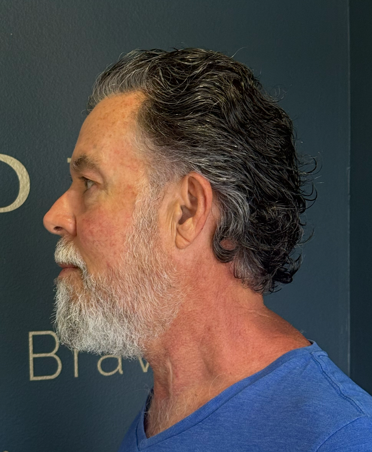 The image shows a man with salt-and-pepper hair and beard, wearing a blue t-shirt, standing against a dark background.