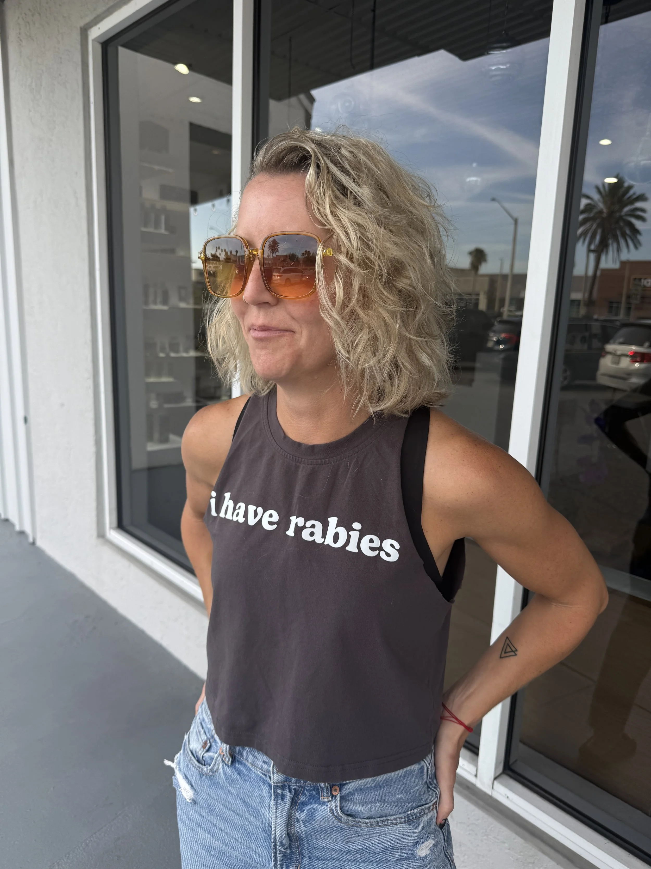 Close-up of a woman with curly blonde hair wearing dark tank top, denim shorts, and orange sunglasses with a reflection of palm trees, standing outside near a glass storefront with palm trees and parked cars in the background.