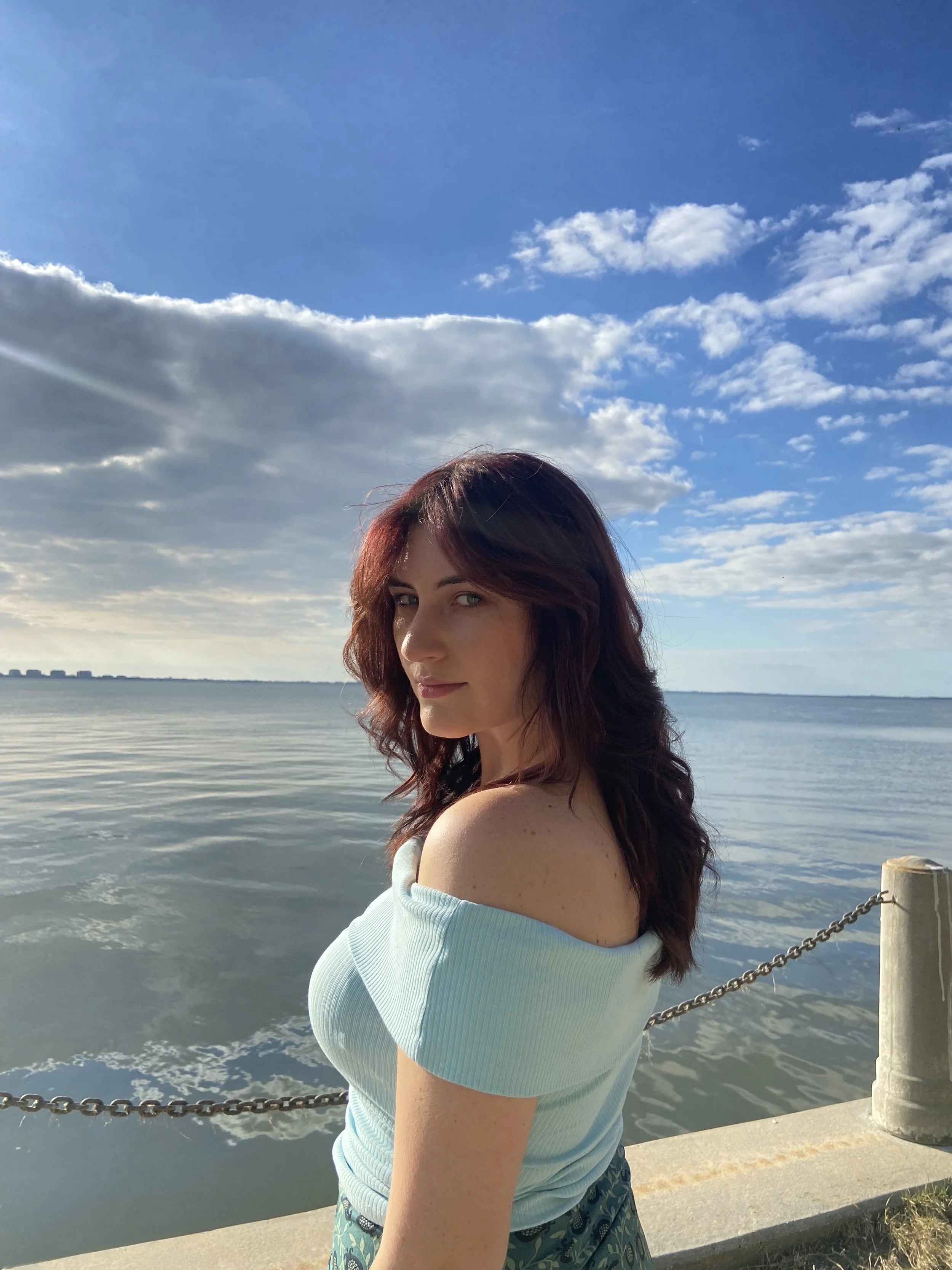 A woman with reddish-brown hair standing near a body of water during daytime, with a partly cloudy sky overhead. She is wearing a light blue off-the-shoulder top and a patterned skirt, and is looking at the camera with a slight smile.