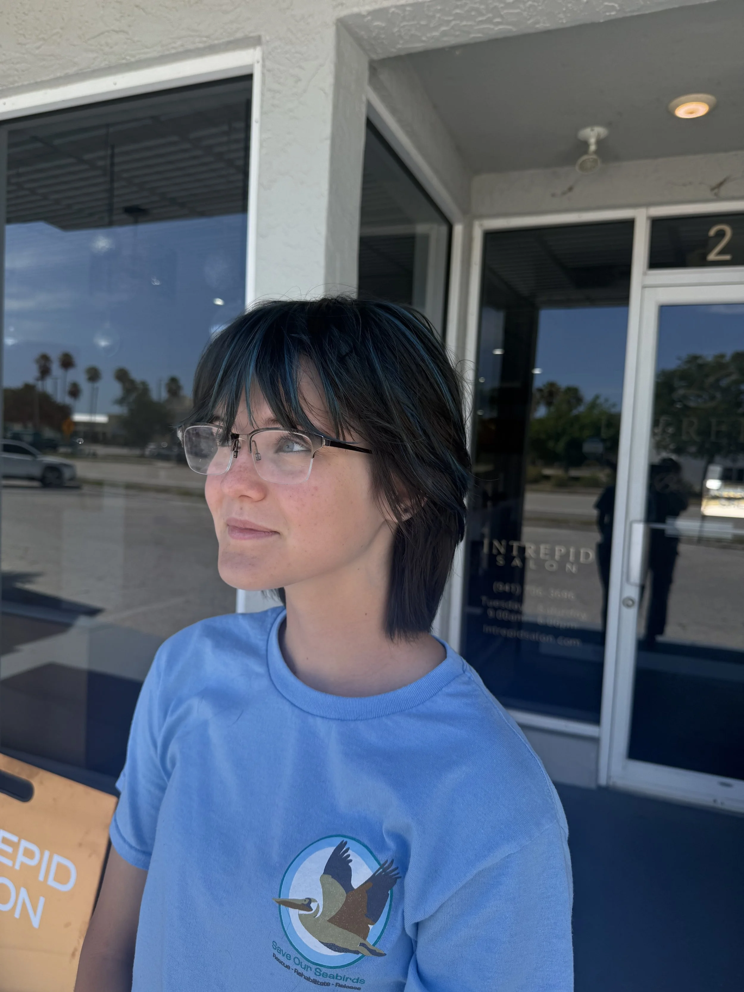 A person with black hair and glasses wearing a blue T-shirt with a logo of a duck and the text "Save Our Seabirds" standing outside a building with glass doors and windows.