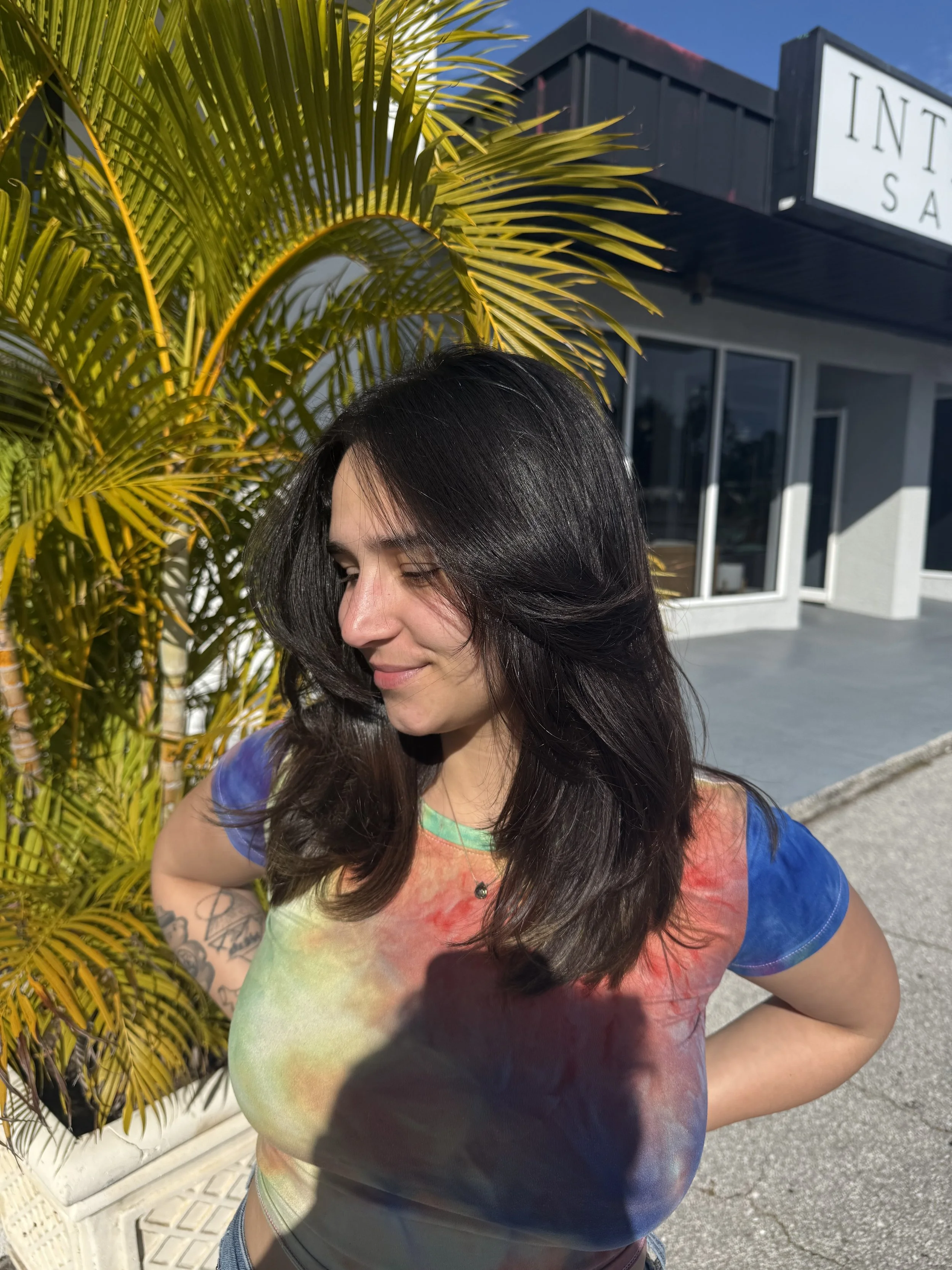 A woman with dark, wavy hair and tattoos on her arm, wearing a colorful tie-dye shirt, standing outdoors next to a palm plant with a building in the background. The building has a sign that reads 'INTER... S'.
