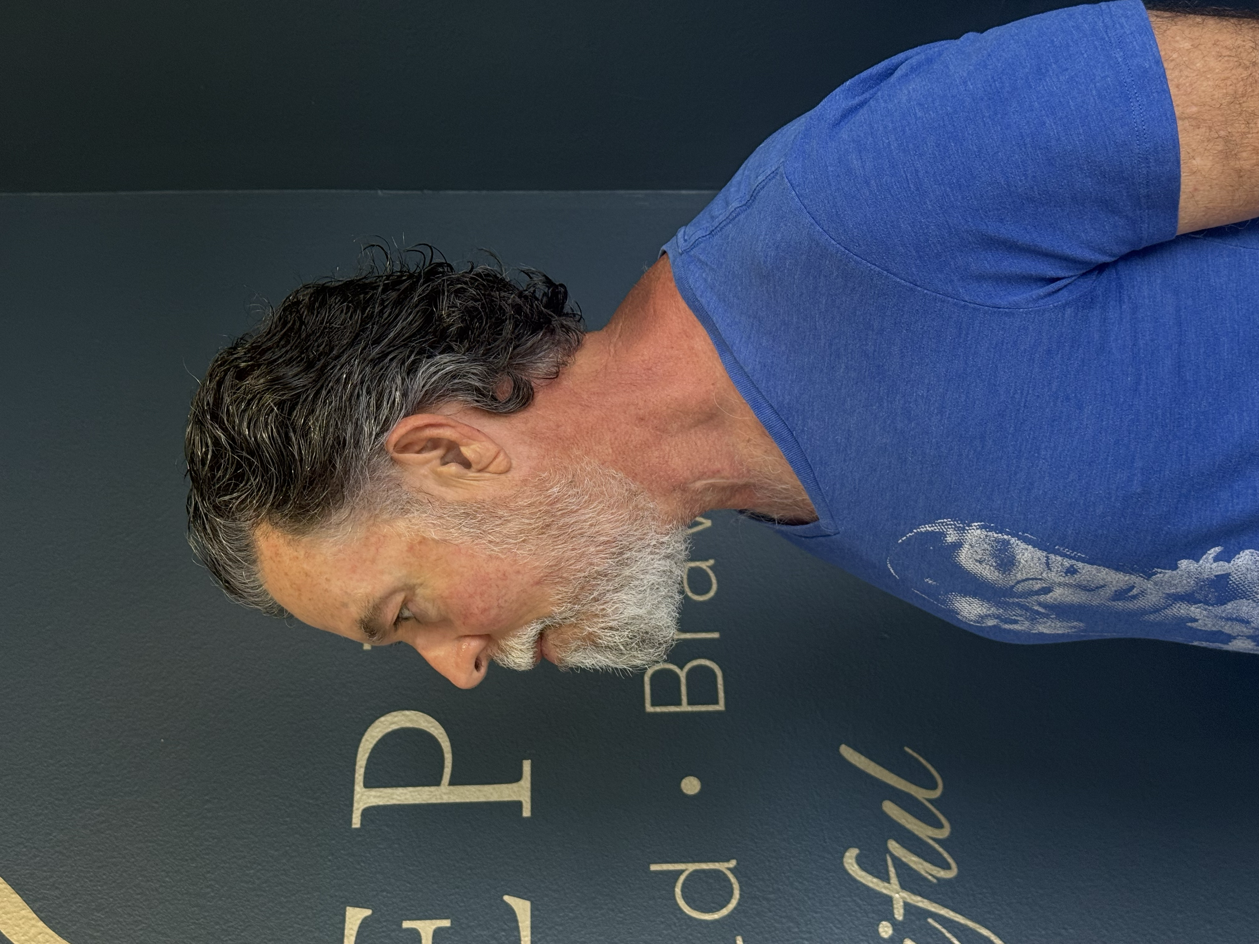 The image shows a man with salt-and-pepper hair and beard, wearing a blue t-shirt, standing against a dark background.