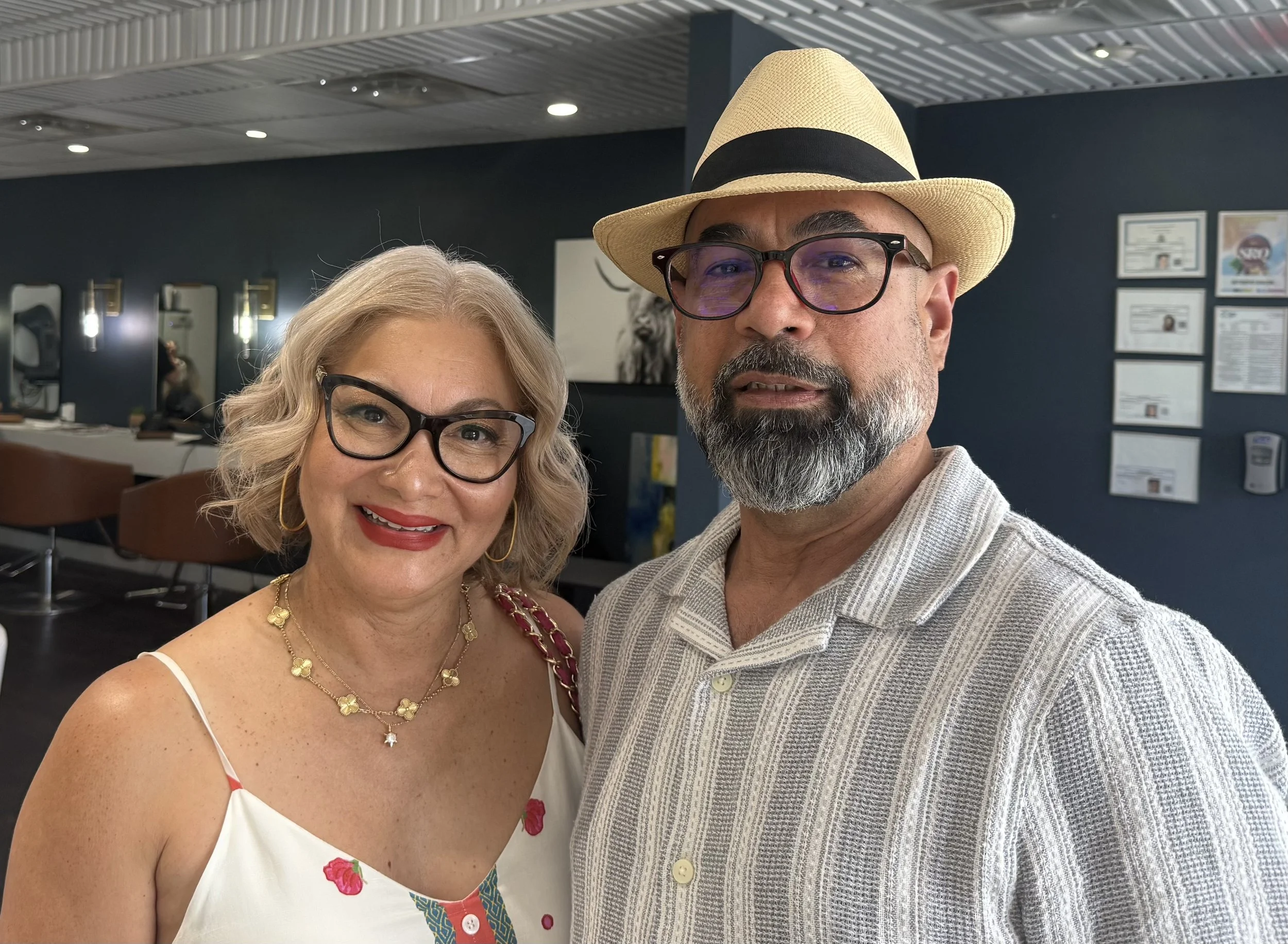 A smiling woman with blonde hair, glasses, and jewelry standing next to a man with glasses, a beard, wearing a straw hat and a light-colored shirt, in an indoor setting with framed pictures and certificates on the wall.