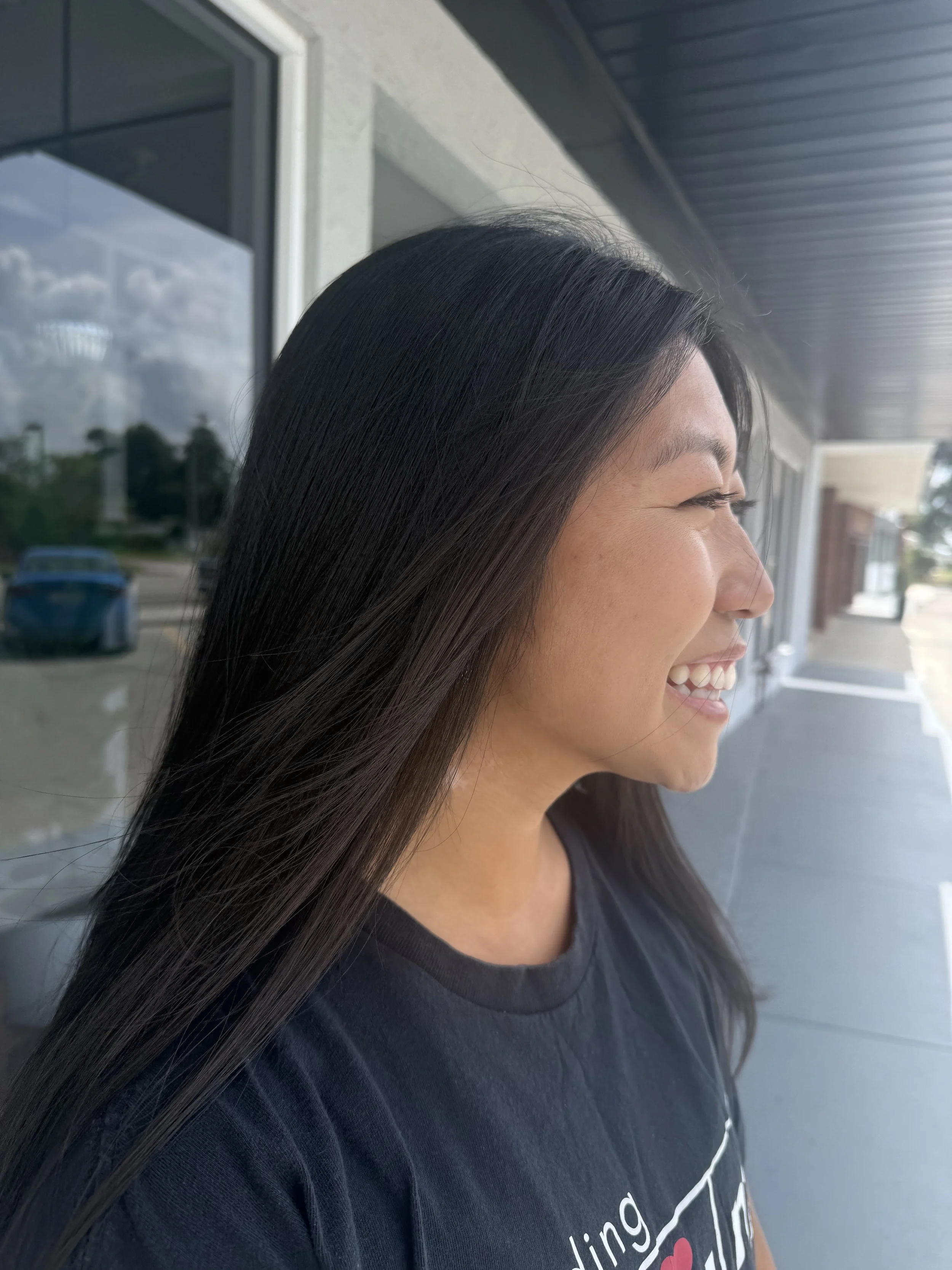 A woman with long black hair smiling outside near a building with large windows and a covered porch.
