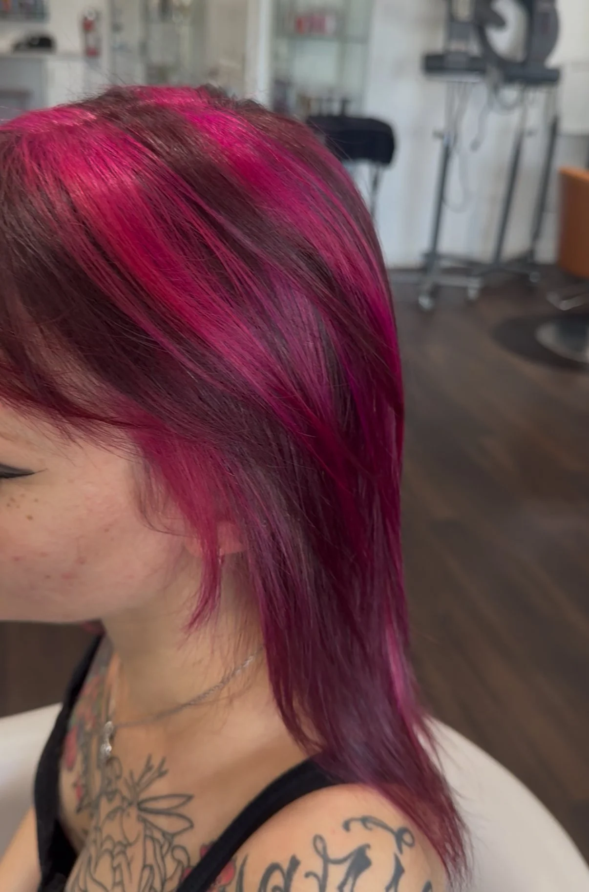 A person with short, vibrant pink hair and tattoos on their shoulder and chest, in a salon or hair studio setting.