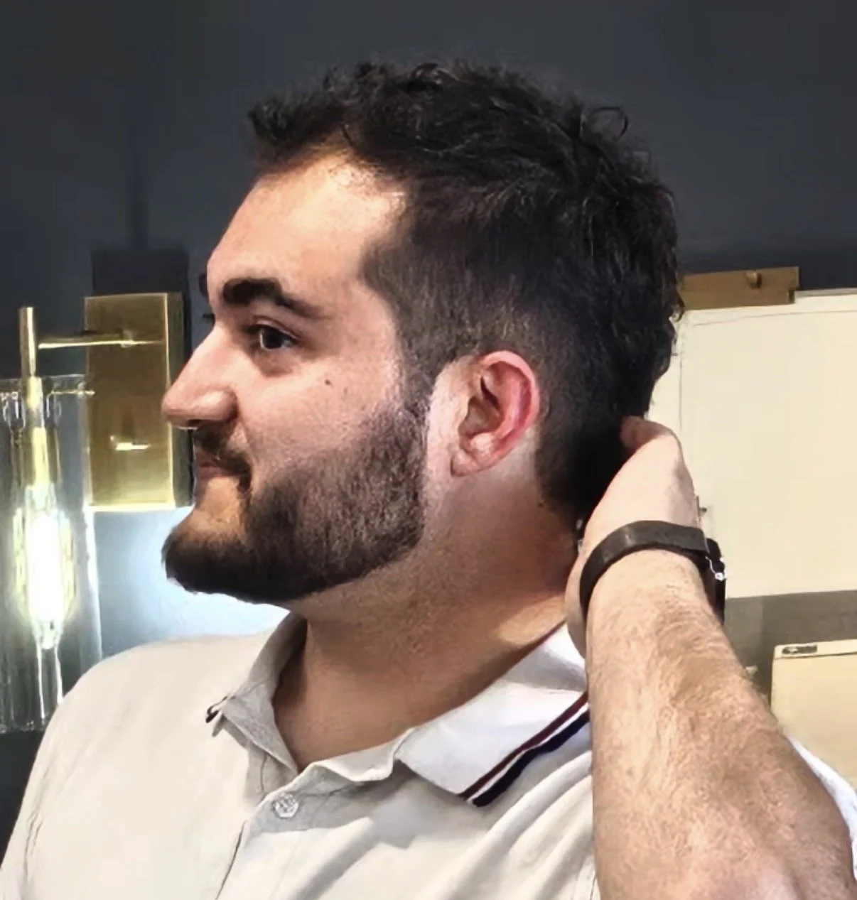 A man with dark, curly hair and a beard is touching the back of his neck. He is wearing a gray collared shirt and a black watch. The background has industrial-style decor with metallic and glass elements.