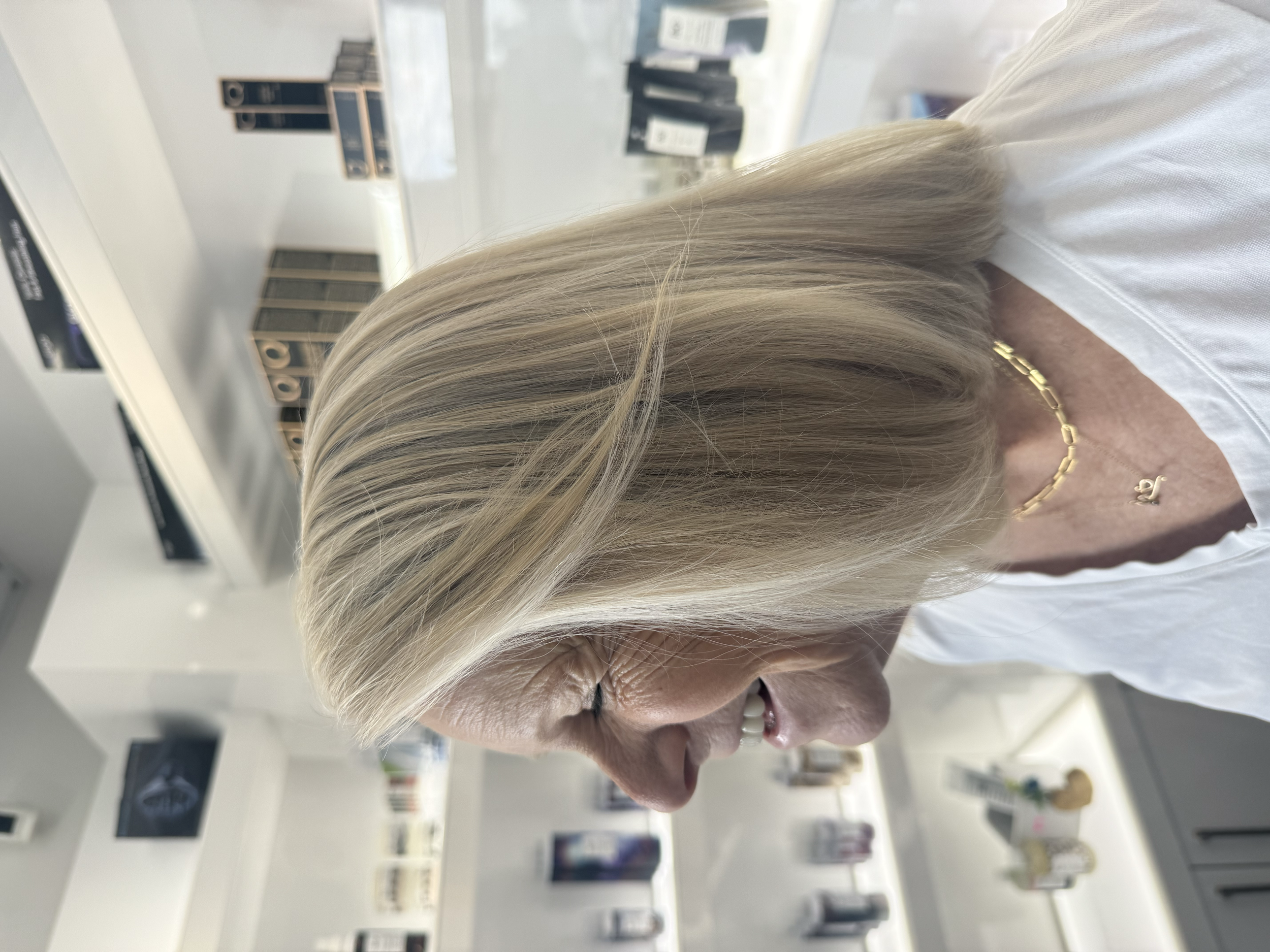 Close-up of elderly woman with light blonde hair, wearing a gold necklace, smiling, in a store or library with shelves and black packages in the background.
