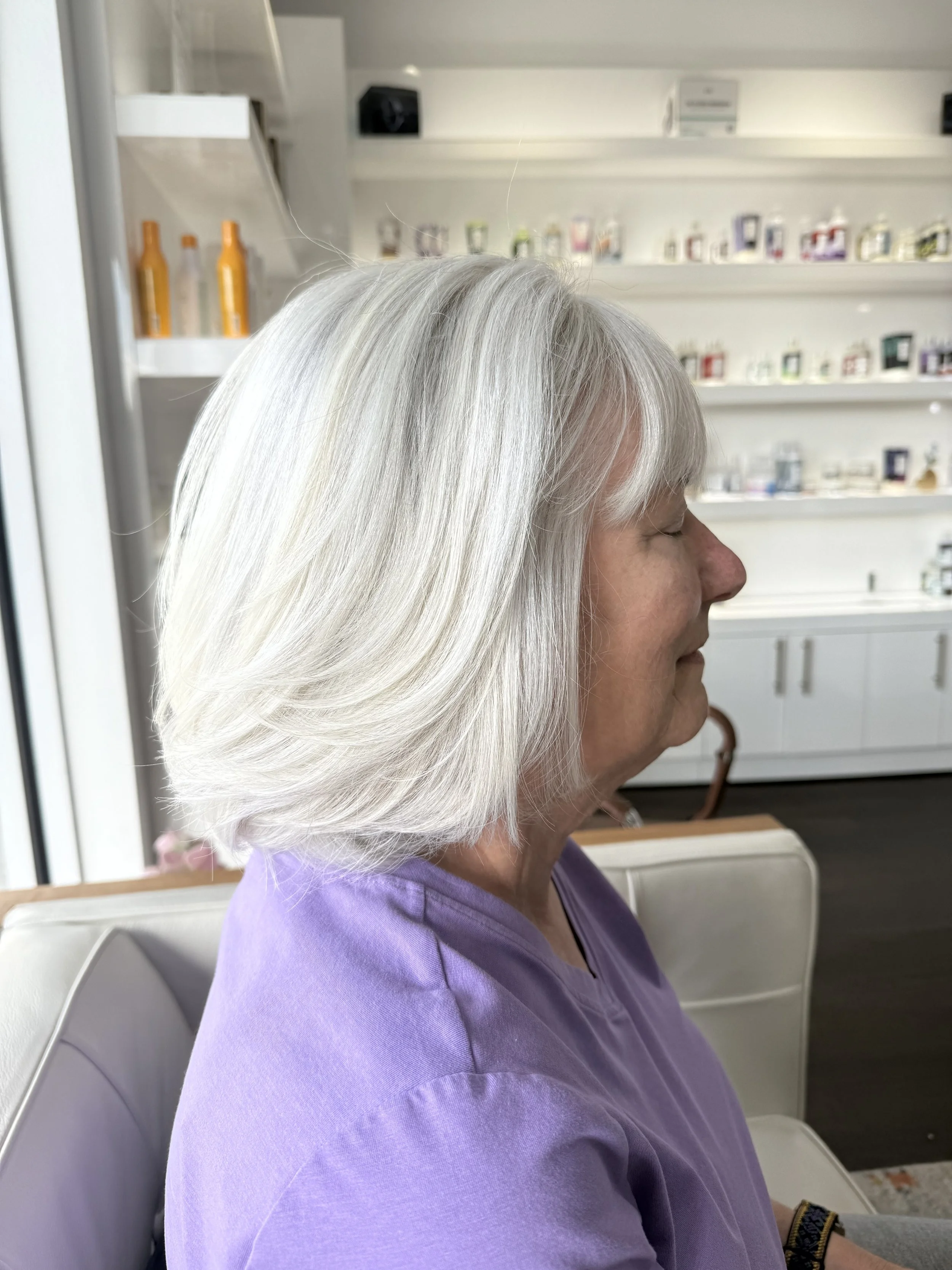 Profile of an elderly woman with white hair sitting with her eyes closed in a waiting room or salon, wearing a lavender top.