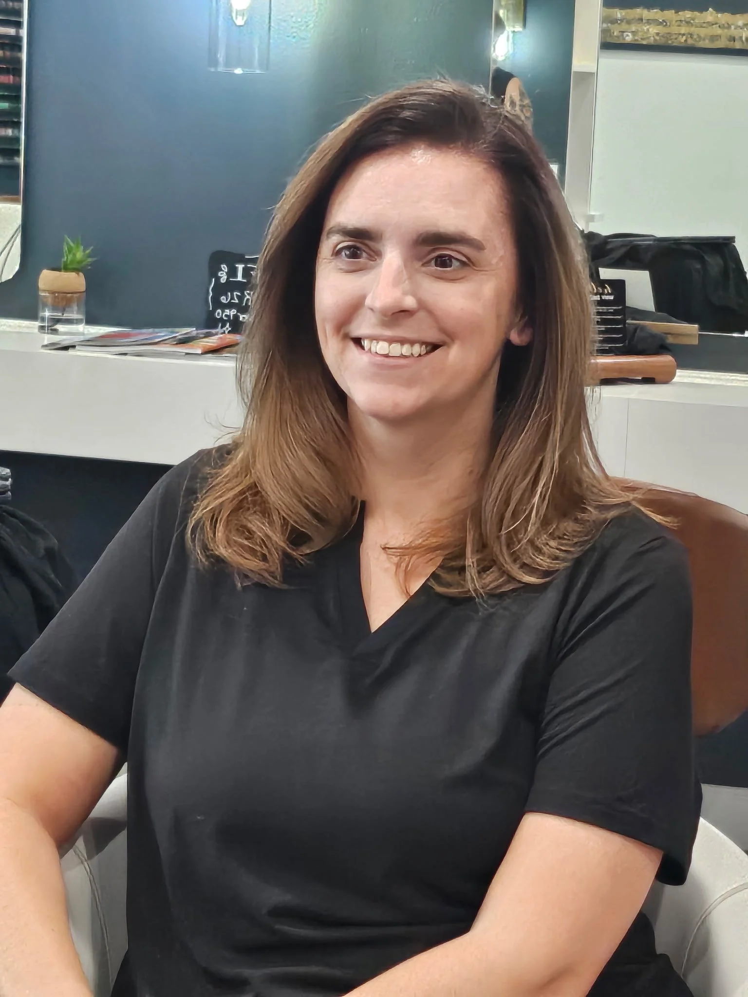 A woman with shoulder-length brown hair sitting in a casual indoor setting, smiling, wearing a black shirt.