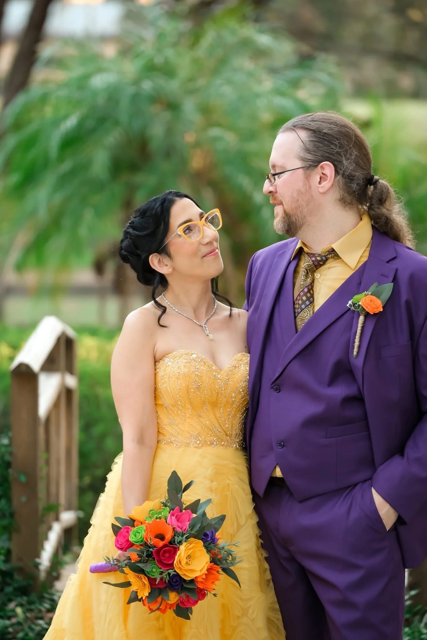 A bride and groom standing together outdoors, the bride in a yellow gown and the groom in a purple suit, smiling at each other, with a colorful bouquet of flowers.