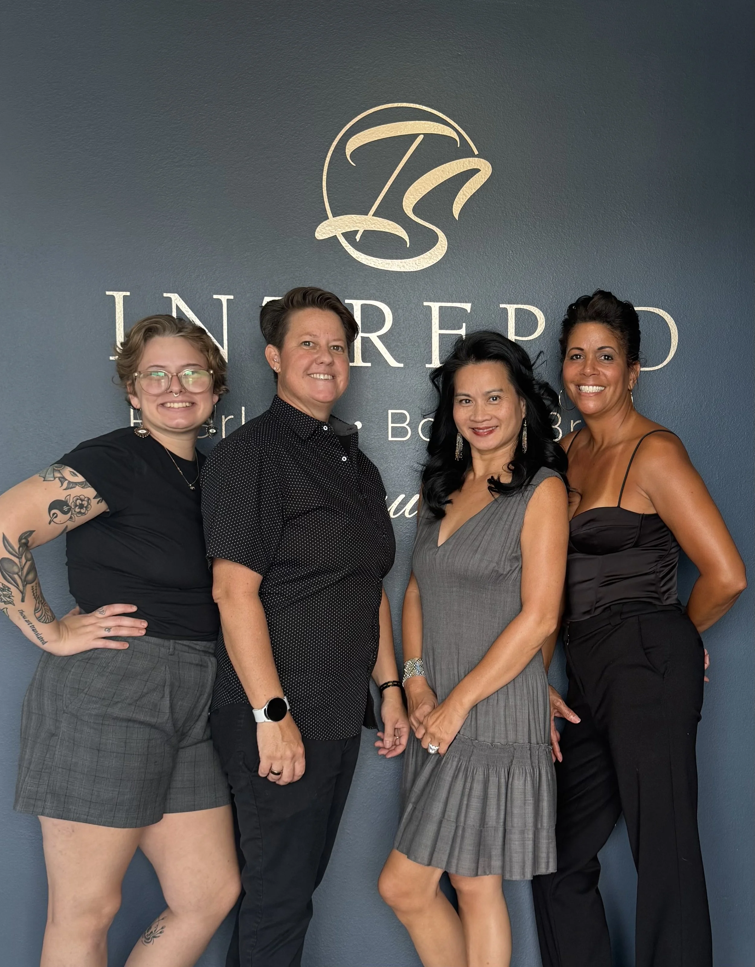 Four women standing together in front of a dark gray wall with silver writing and logo. They are smiling and dressed in fashionable attire, with various hairstyles and accessories.