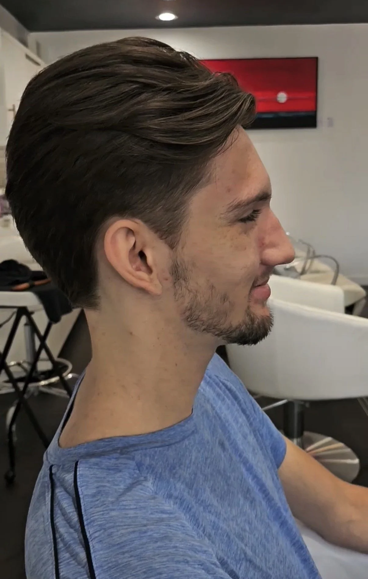 Side profile of a young man with styled brown hair, a slight beard, wearing a blue t-shirt, sitting in a hair salon.