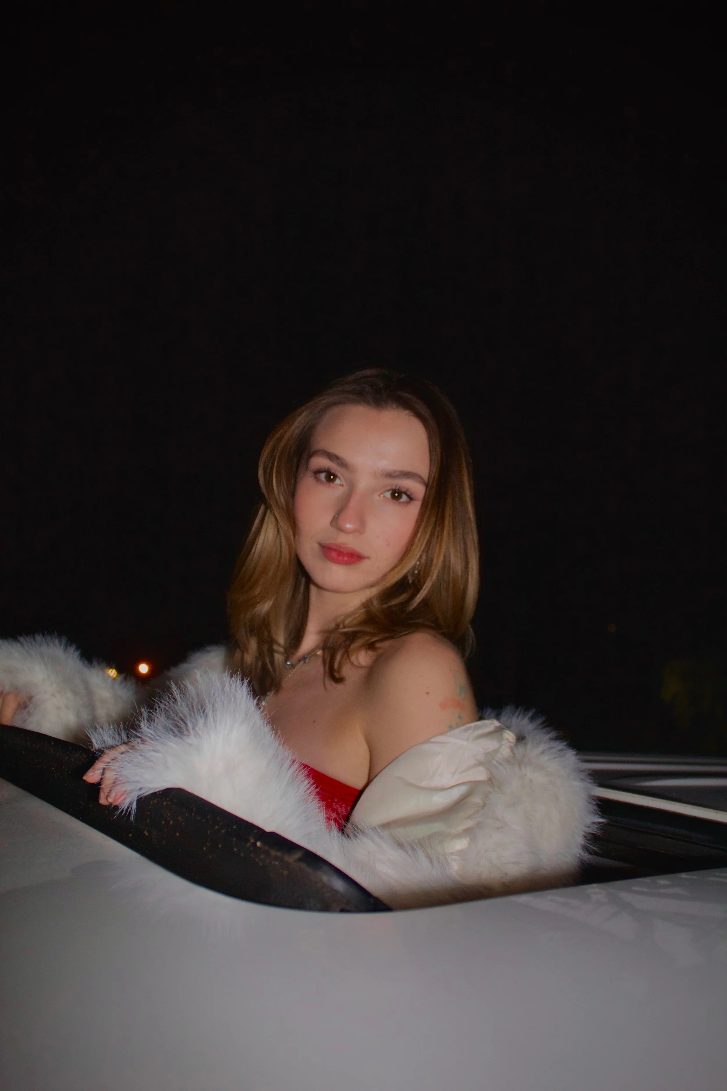 Young woman with long brown hair looking at the camera, wearing a white fur coat and a red dress, sitting in a car at night with a dark sky above.