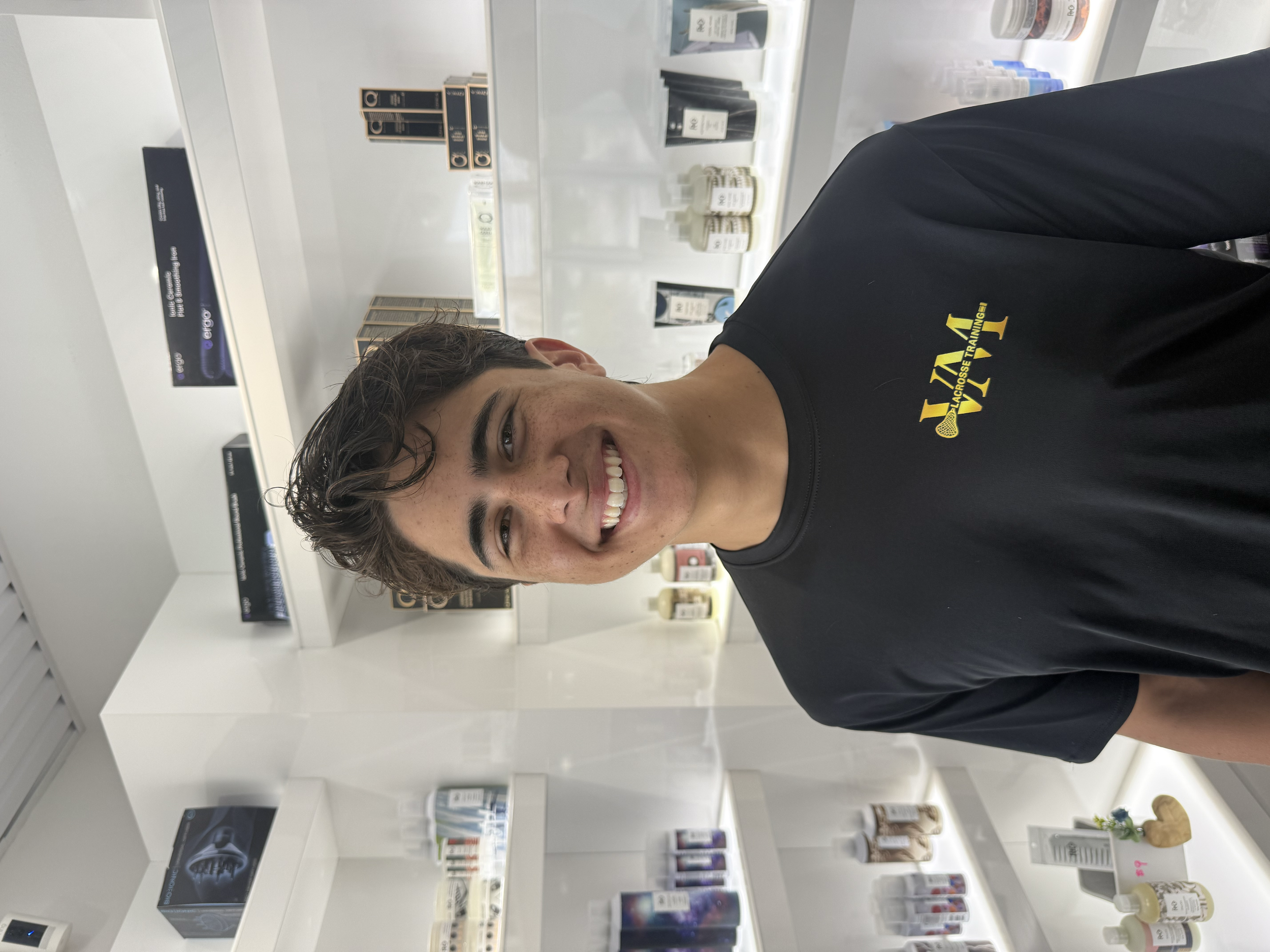 A young man with brown wavy hair and light skin smiling and looking at the camera while shopping for skincare and beauty products in a store. He is wearing a black t-shirt with yellow text that reads 'ALL CROSS TRAINING'. The store has bright white s
