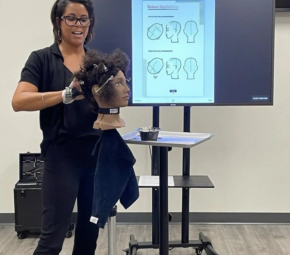 A hairstylist demonstrating relaxation techniques on a mannequin head with styled hair, using diagrams on a large screen behind her to show head positions.