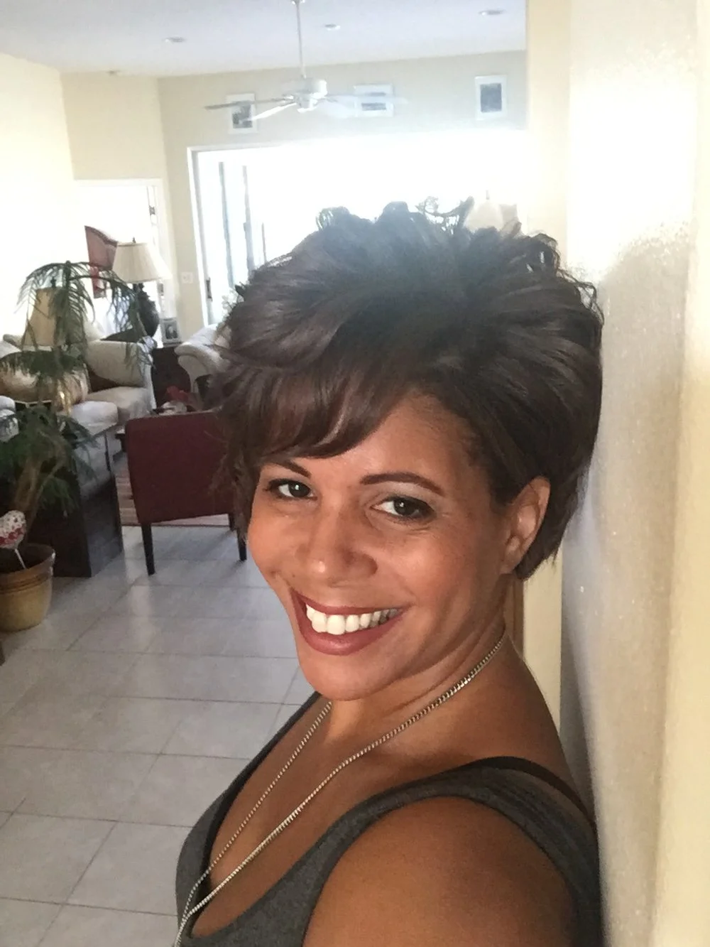 A woman with short, layered dark hair smiling and taking a selfie indoors with a living room background.