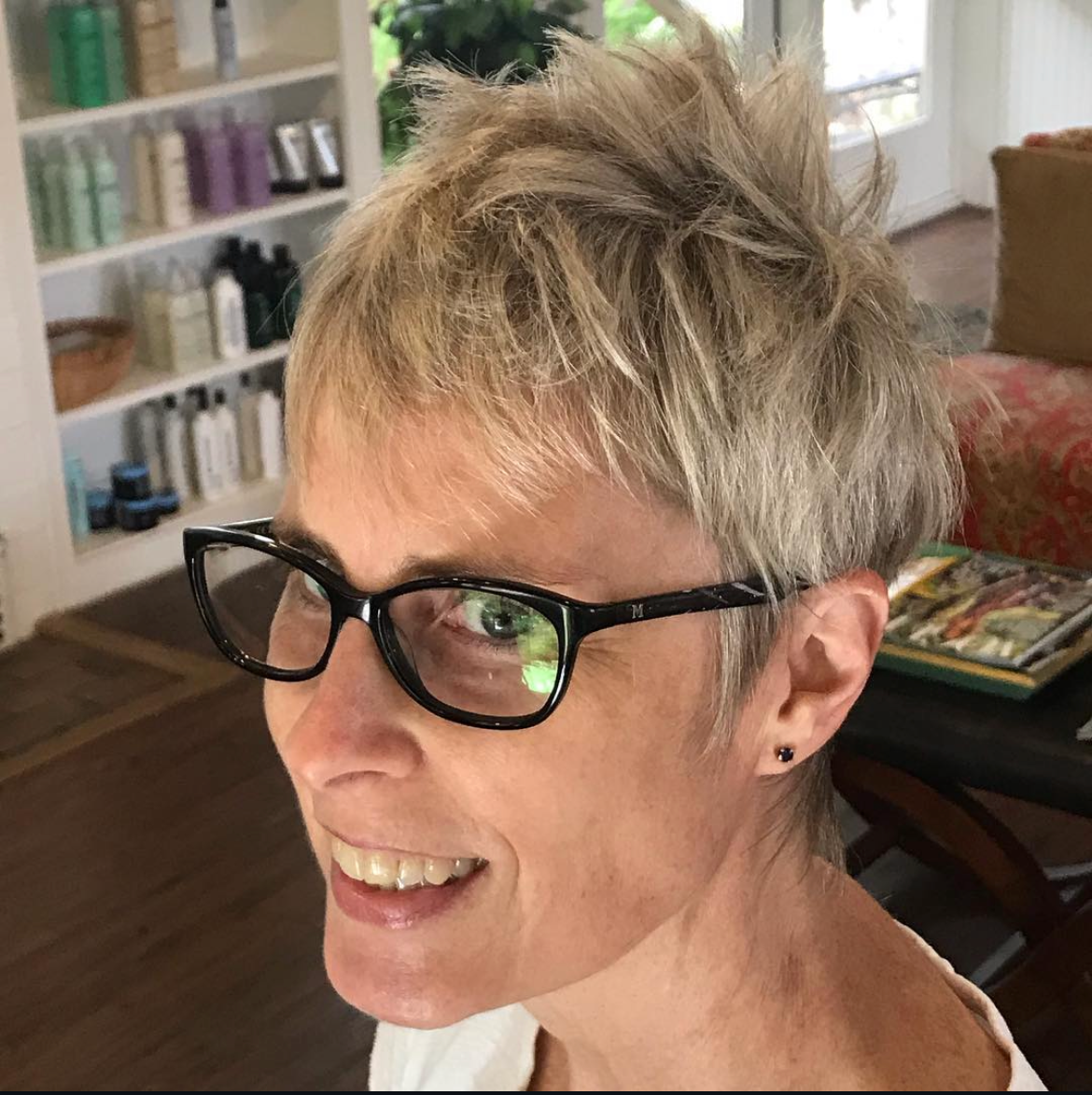Close-up of a woman with short blonde hair, wearing black glasses, smiling, in a cozy room with bookshelves and a window in the background.