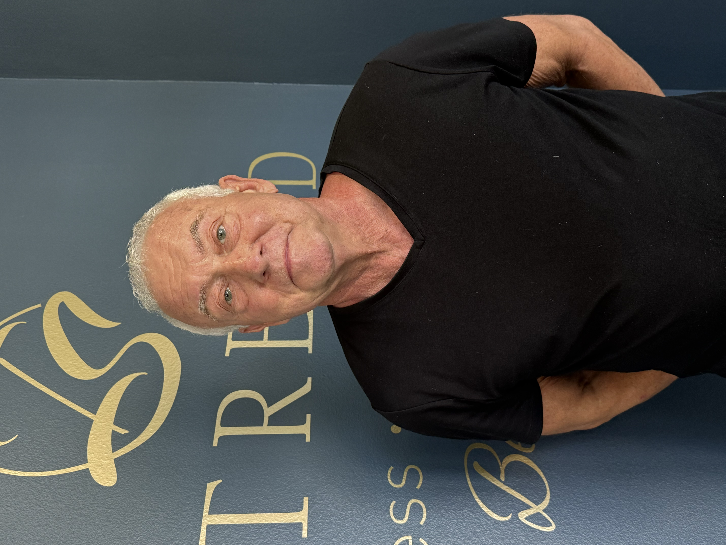 An elderly man with white hair smiling at the camera, wearing a black t-shirt, standing in front of a dark blue wall with gold lettering.