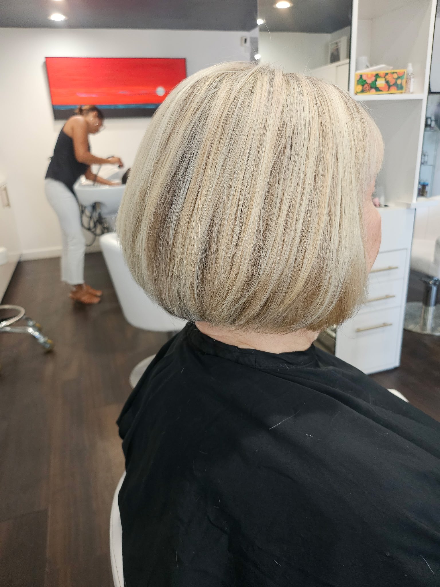 Close-up of a woman with a blonde, straight bob hairstyle in a salon, with another woman washing hair in the background.