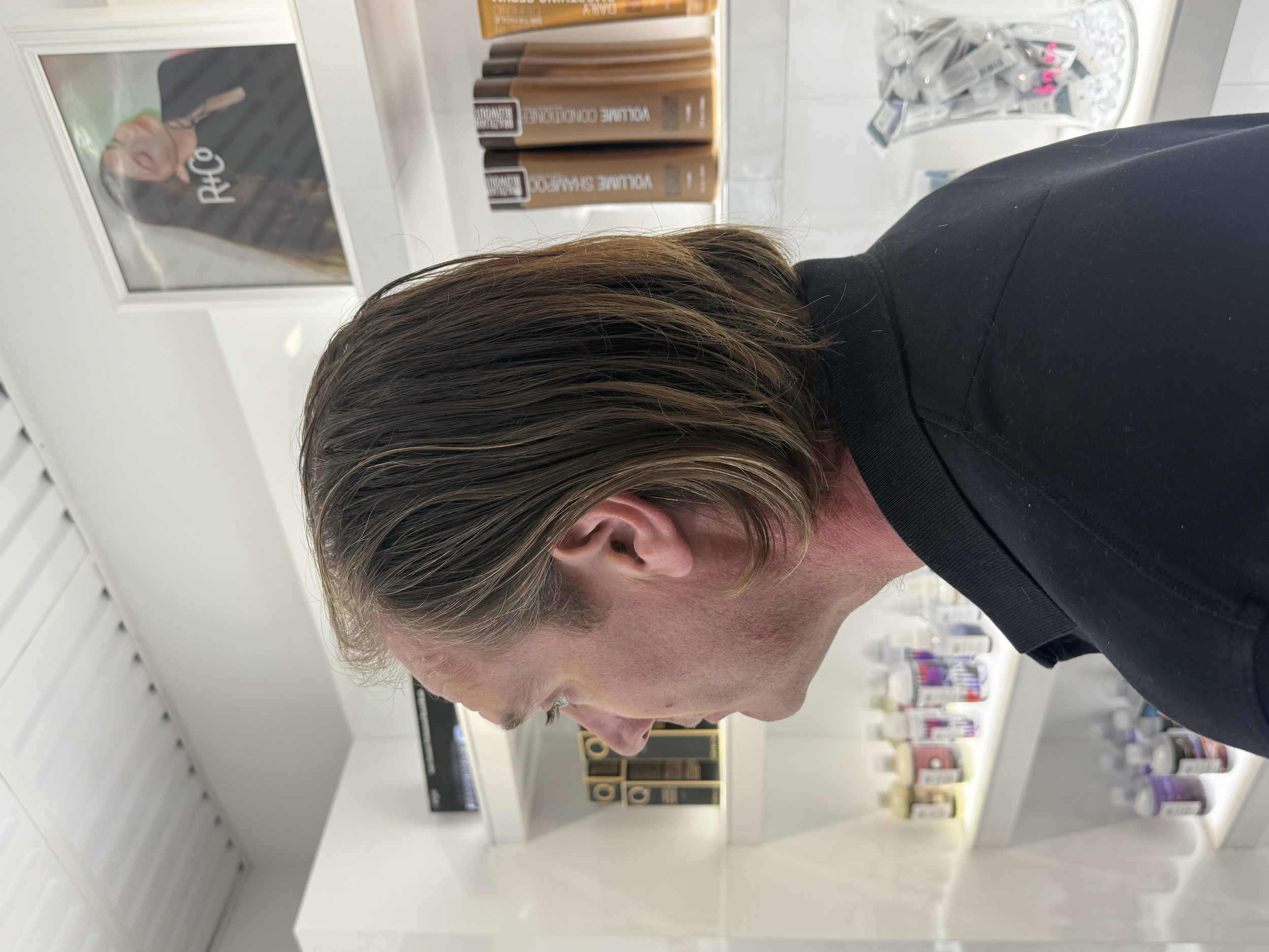 Side profile of a man with brown hair and a mustache, wearing a black shirt, in a store aisle with hair products on the shelves.